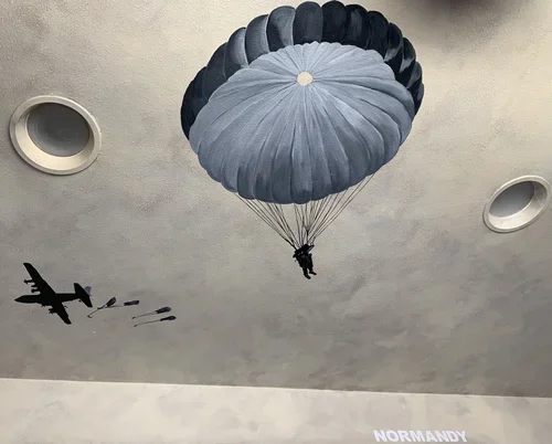 Ceiling mural depicting a paratrooper descending with a parachute and a fighter jet releasing bombs.