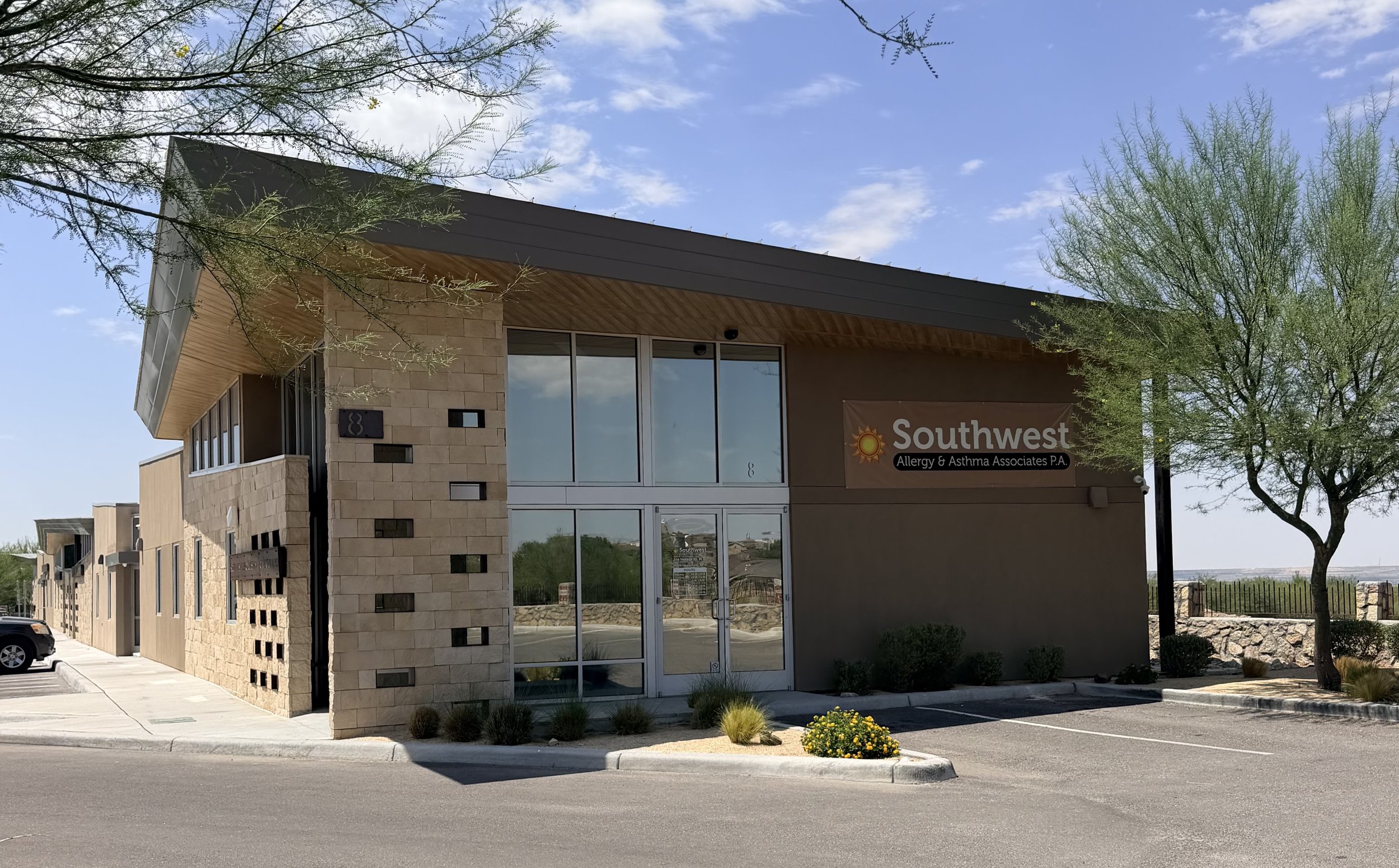 The image shows a modern building with a sign that reads 'Southwest Allergy & Asthma Associates P.A.' under a partly cloudy sky. There are trees and a small landscaped area with flowers and bushes in front of the building, and a parking lot with a few cars.
