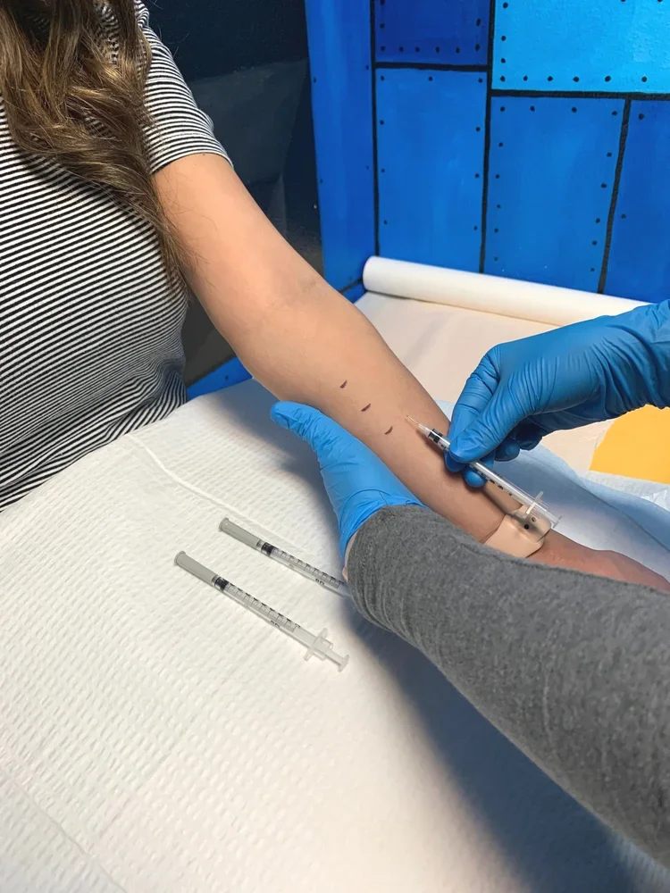 A person with long hair receiving an injection in their forearm from a healthcare professional wearing blue gloves, while the patient is seated at a medical station with two additional syringes on a white paper cover.