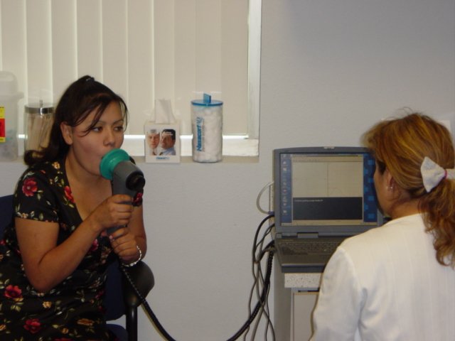 A woman with dark hair in a floral dress is speaking into a medical device, likely an otoscope, while a healthcare professional with red hair tied with a white scrunchie looks at a computer monitor in a clinical setting.