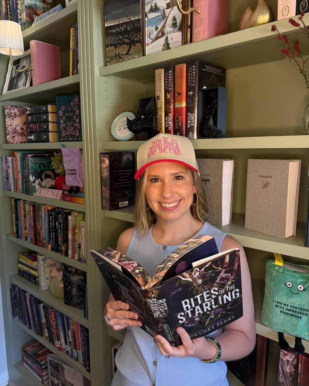 Woman wearing a cap smiling while holding the open book 'Rites of the Starling,' standing in front of a green bookshelf filled with books
