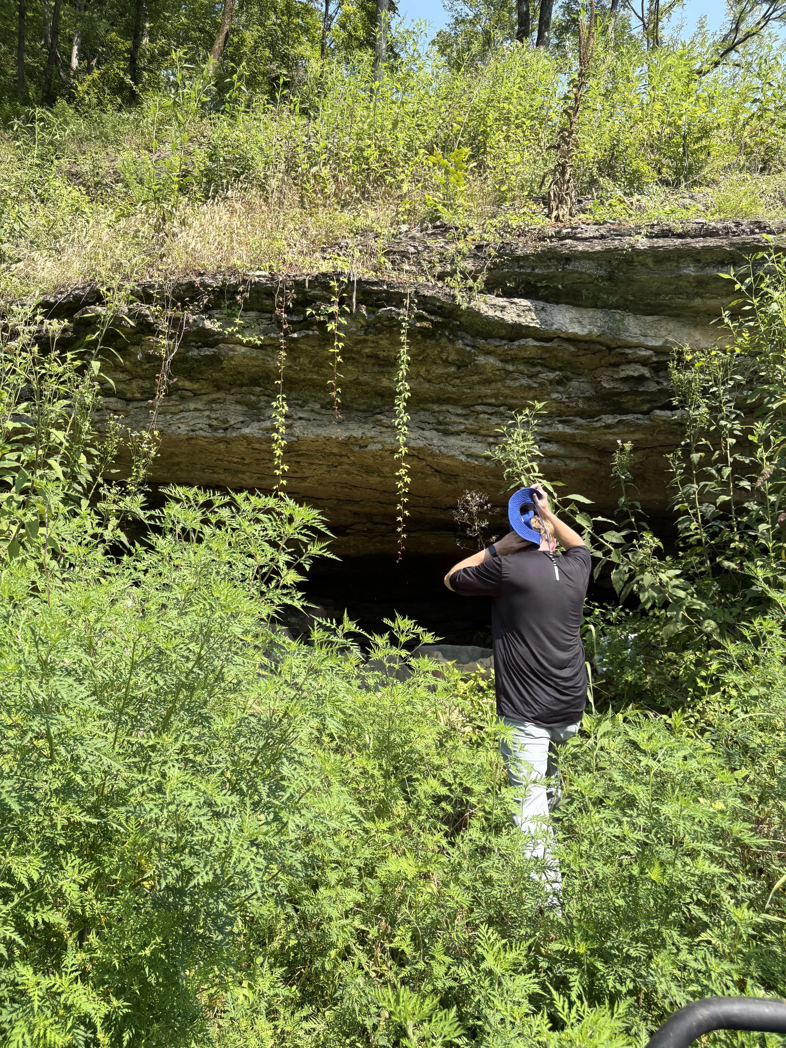 Person wearing a black jacket and white pants taking a photograph of a large, overhanging rock formation with lush green vegetation around it.