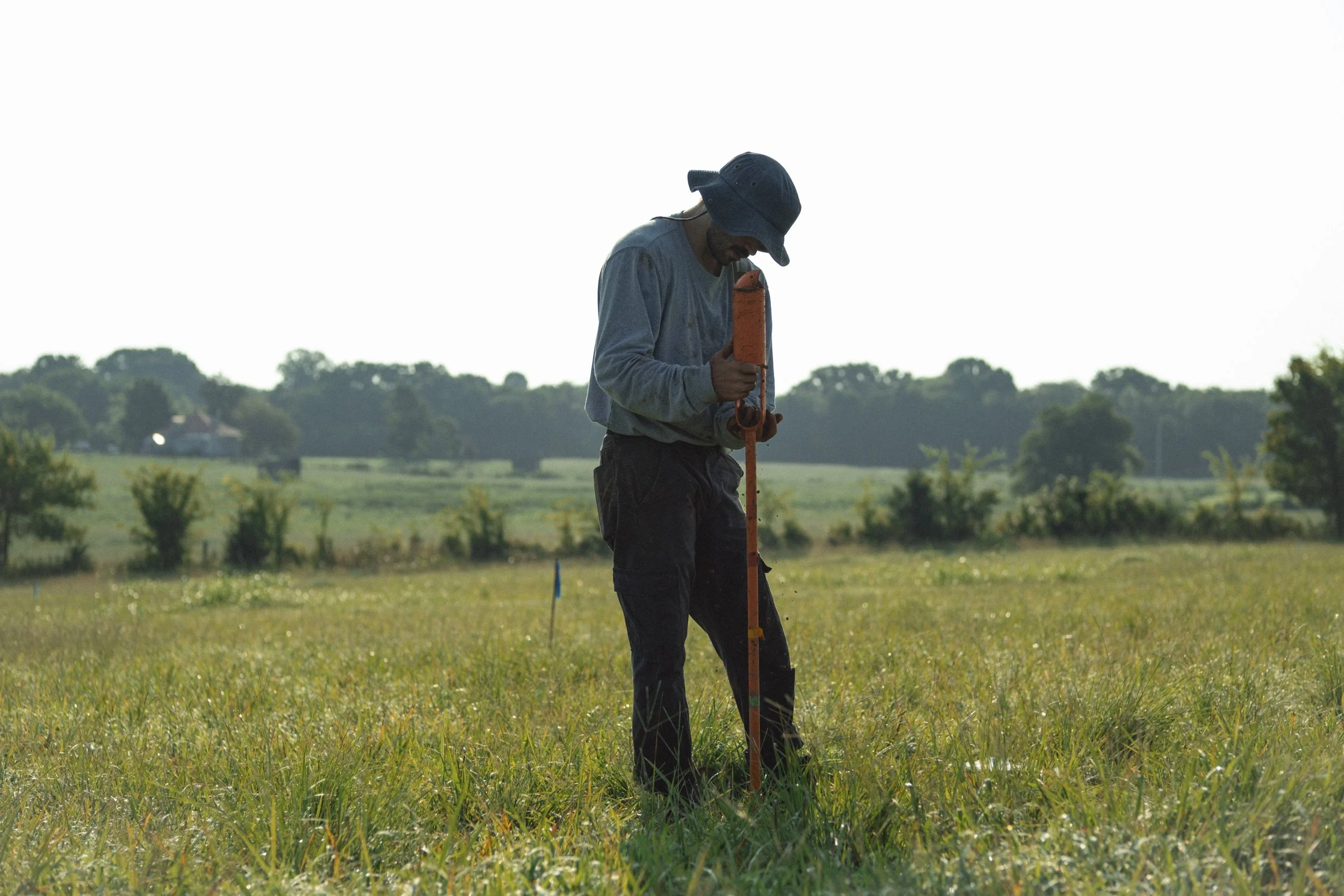 A man wearing a blue bucket hat and a gray long-sleeve shirt is outdoors in a grassy field, holding a measuring rod and looking down at the ground during daytime.