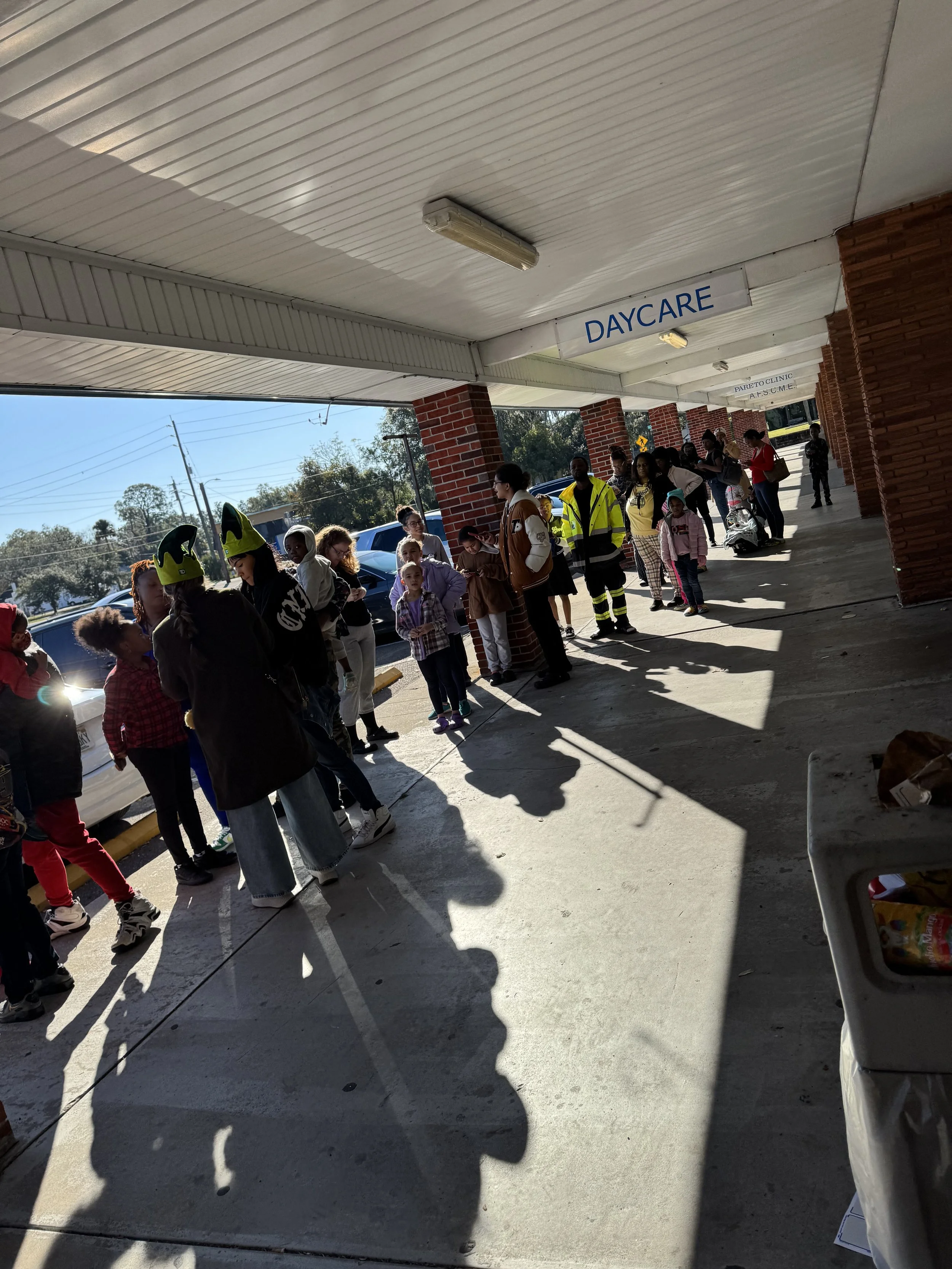 Line of people waiting outside a daycare center on a sidewalk, with some children and adults, including a person wearing a yellow reflective jacket, under a canopy with a sign that says 'DAYCARE'.