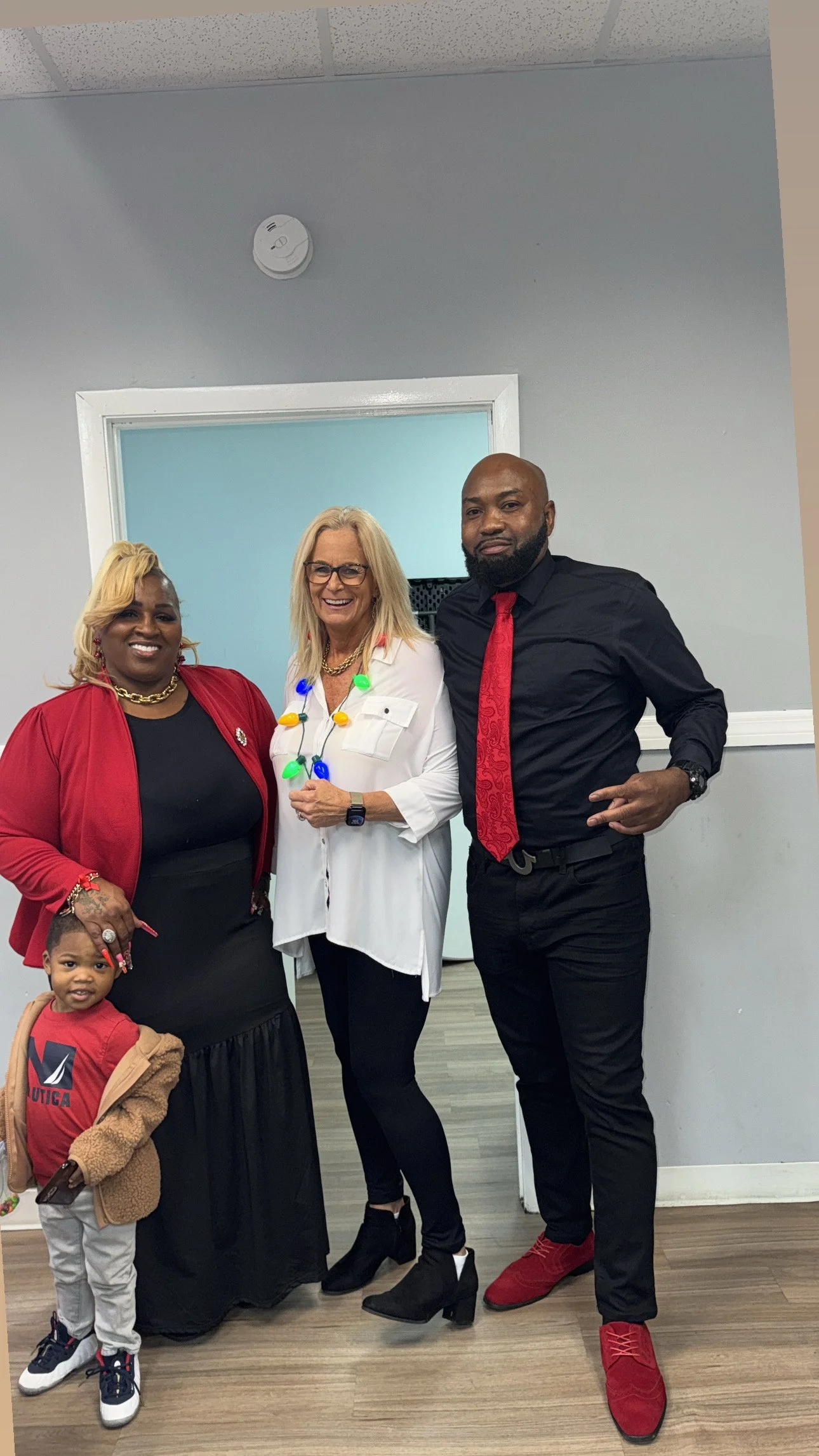 Four people, three adults and one child, standing together inside a room with gray walls. The woman in the middle is wearing glasses, a white shirt with colorful string lights, and is smiling. The woman on the left has blonde hair, glasses, a red jacket, and a black dress. The man on the right has a beard, is dressed in black with a red tie and red shoes. The small boy on the far left is wearing a red shirt, beige jacket, and white sneakers, holding a toy.