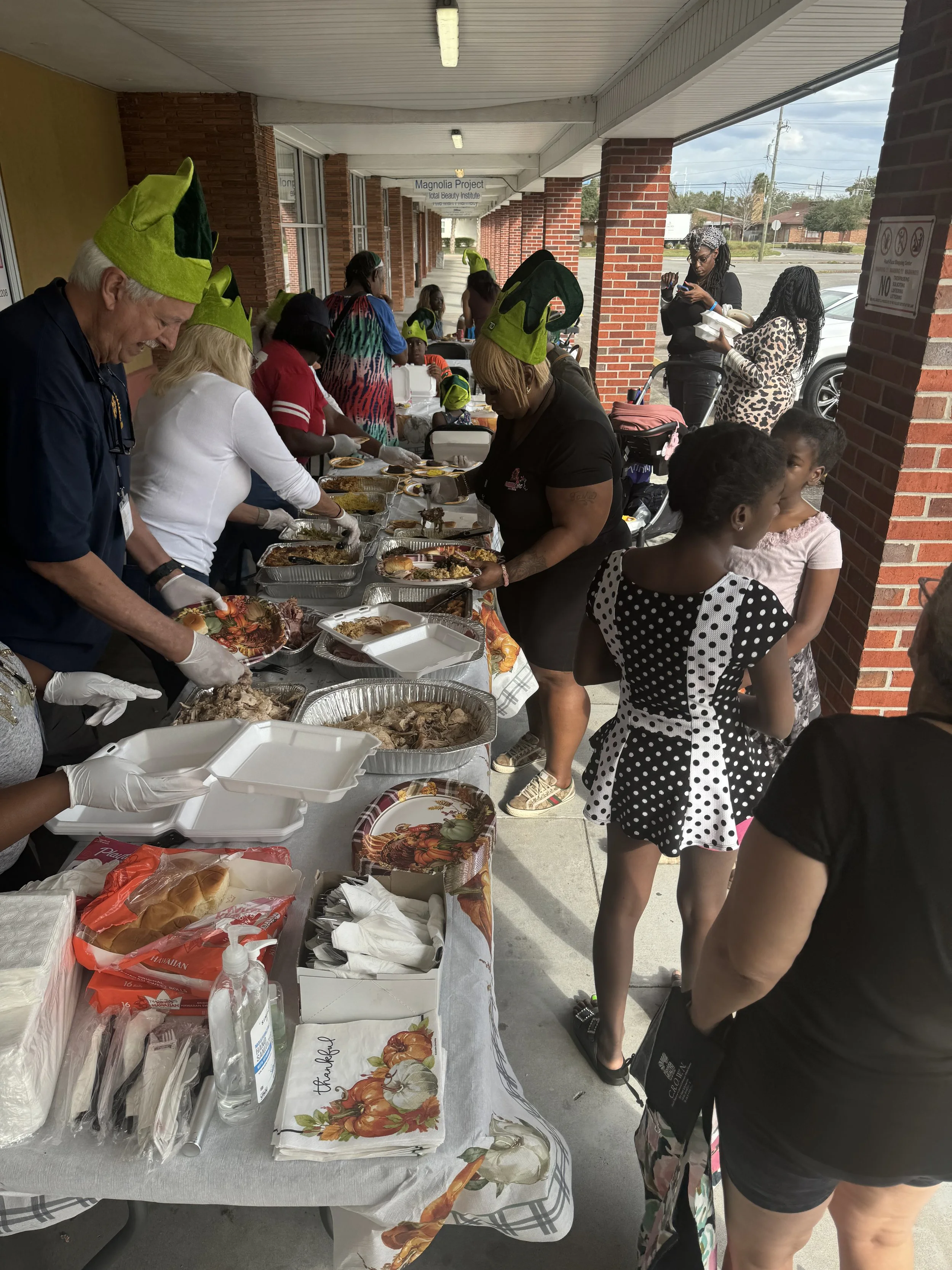 People serving and receiving food at a community event outdoors, with a table filled with trays of food, and individuals in line for a meal, some wearing green elf hats.