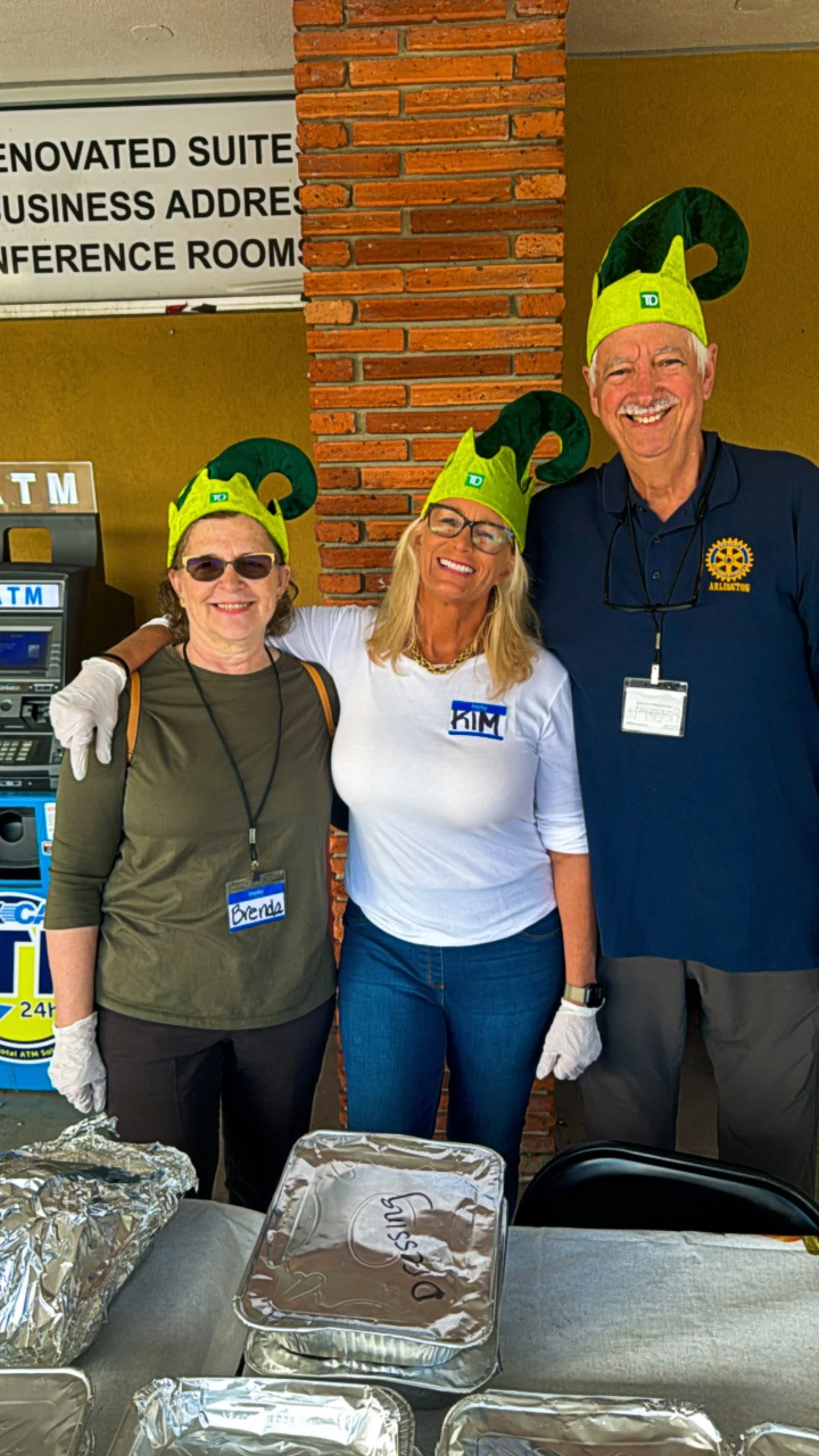 Three people, two women and one man, are standing together at an event. They are wearing elf hats and name tags, with food containers in front of them, likely at a community service event.