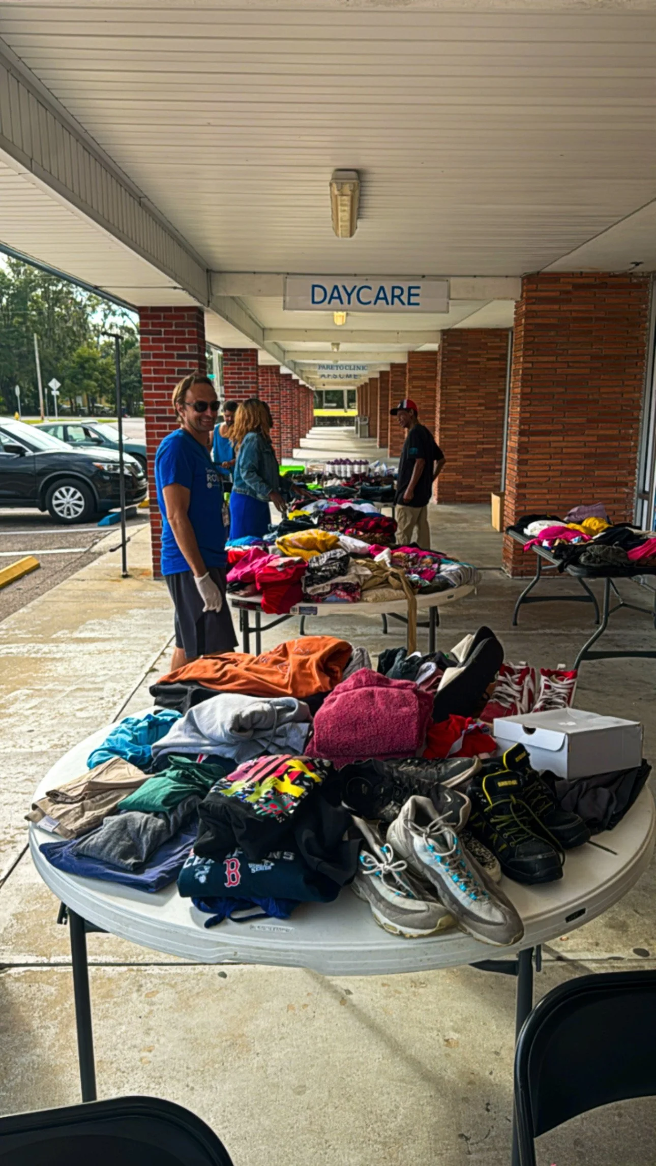 Outdoor table with various clothing, shoes, and accessories for sale at a yard sale outside a building labeled 'DAYCARE' with a parking lot and cars in the background.