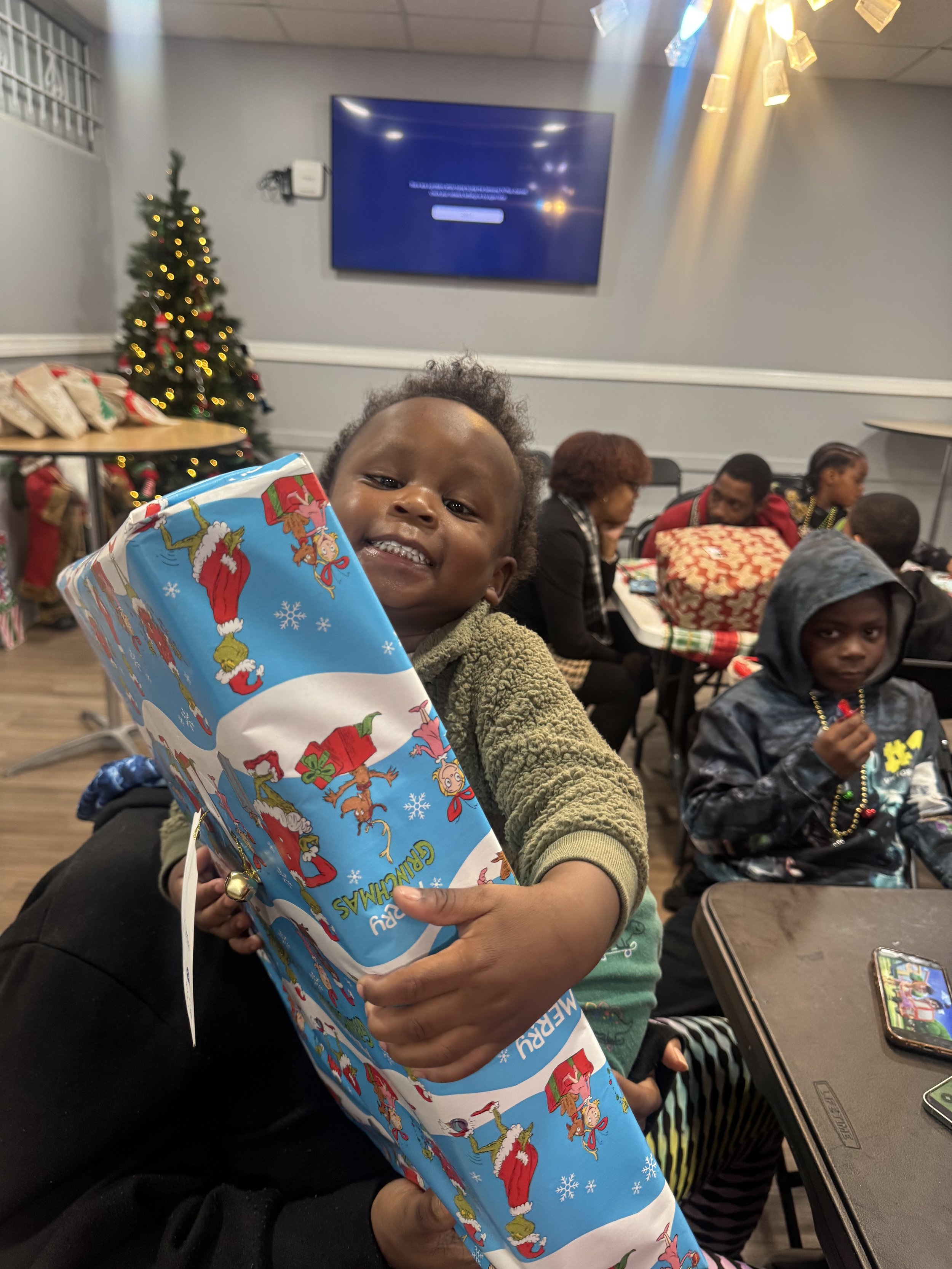 A smiling young boy holding a Christmas-themed gift bag, sitting in a room decorated for Christmas with a Christmas tree and wrapped presents in the background. Other children and adults are seated at tables.
