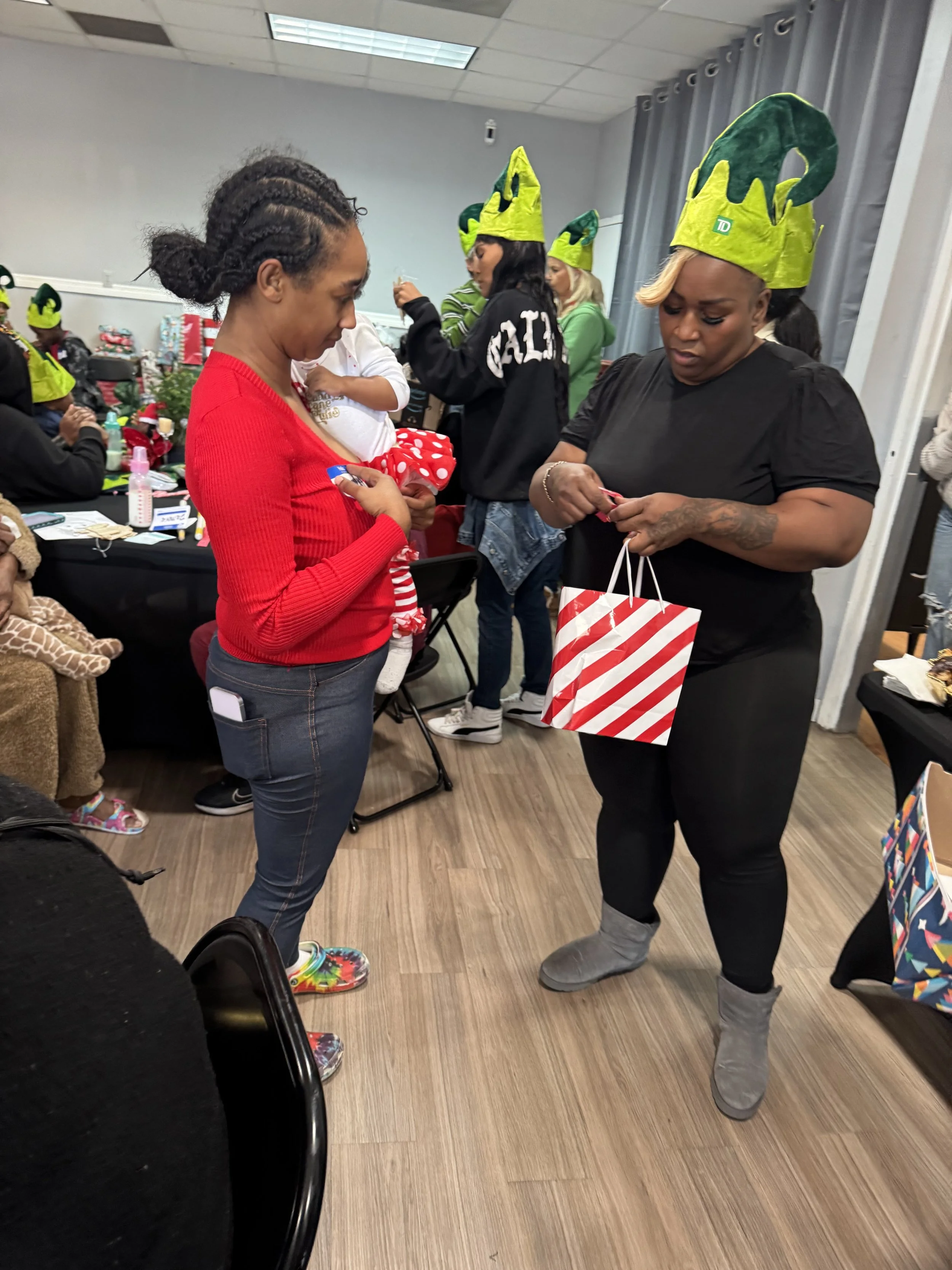 Two women with Christmas gift bags exchanging gifts at a holiday event. Both women are wearing winter boots, and some people in the background are also wearing Christmas hats.