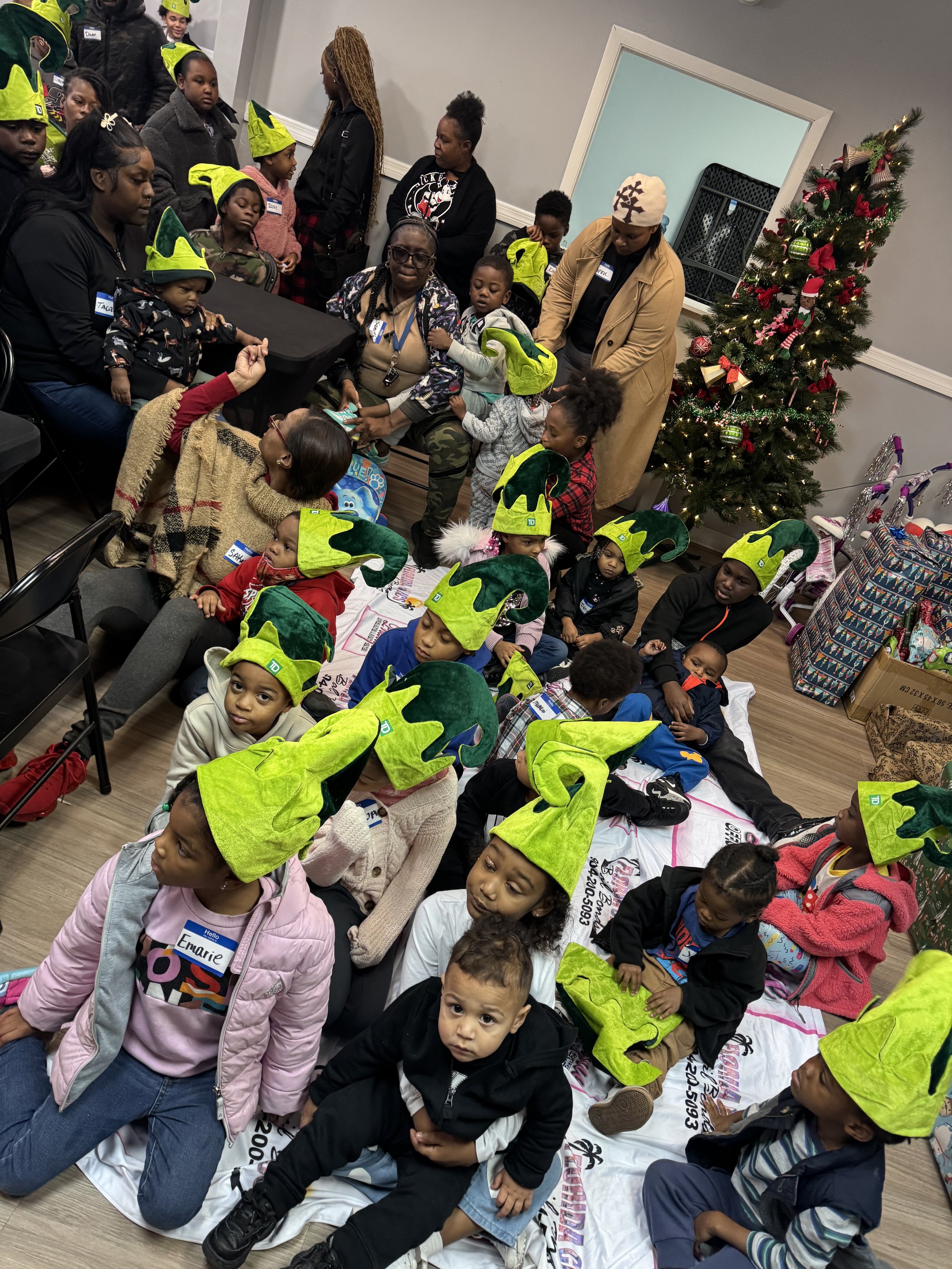 Group of children and adults gathered indoors, with some children wearing bright green elf hats, indicating a holiday event. There is a decorated Christmas tree on the right side of the image, and children are seated on the floor and standing around a table, appearing engaged in an activity or discussion.