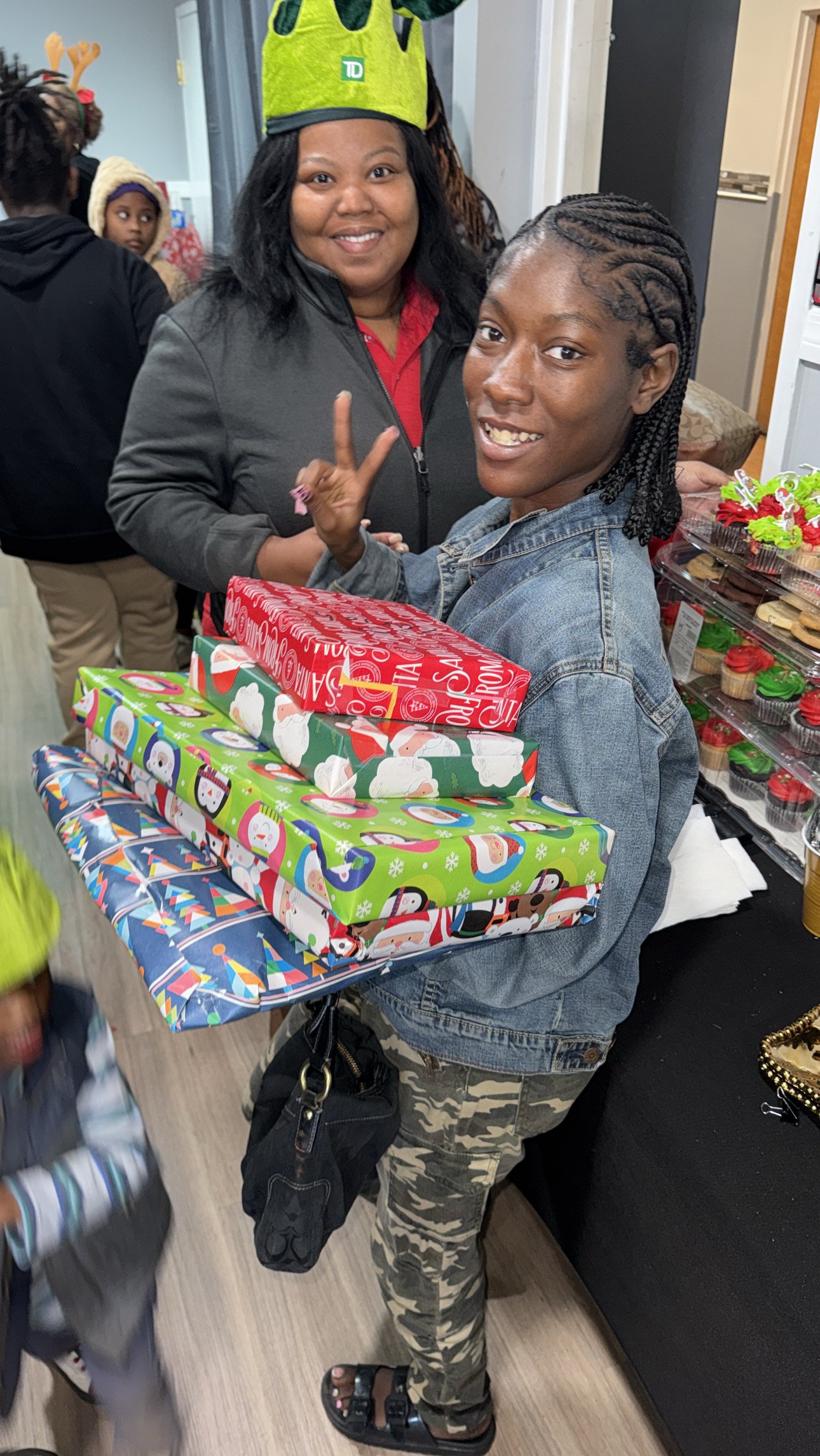 A woman smiling and holding a tray of Christmas presents, with a young person next to her. The presents are decorated with festive wrapping paper, and there are more holiday treats in the background.