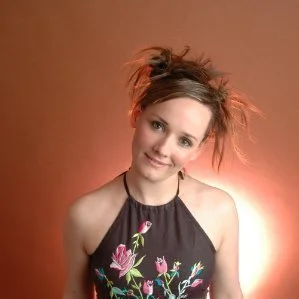 Young woman with messy hair wearing a black floral top, smiling slightly against a warm gradient background.