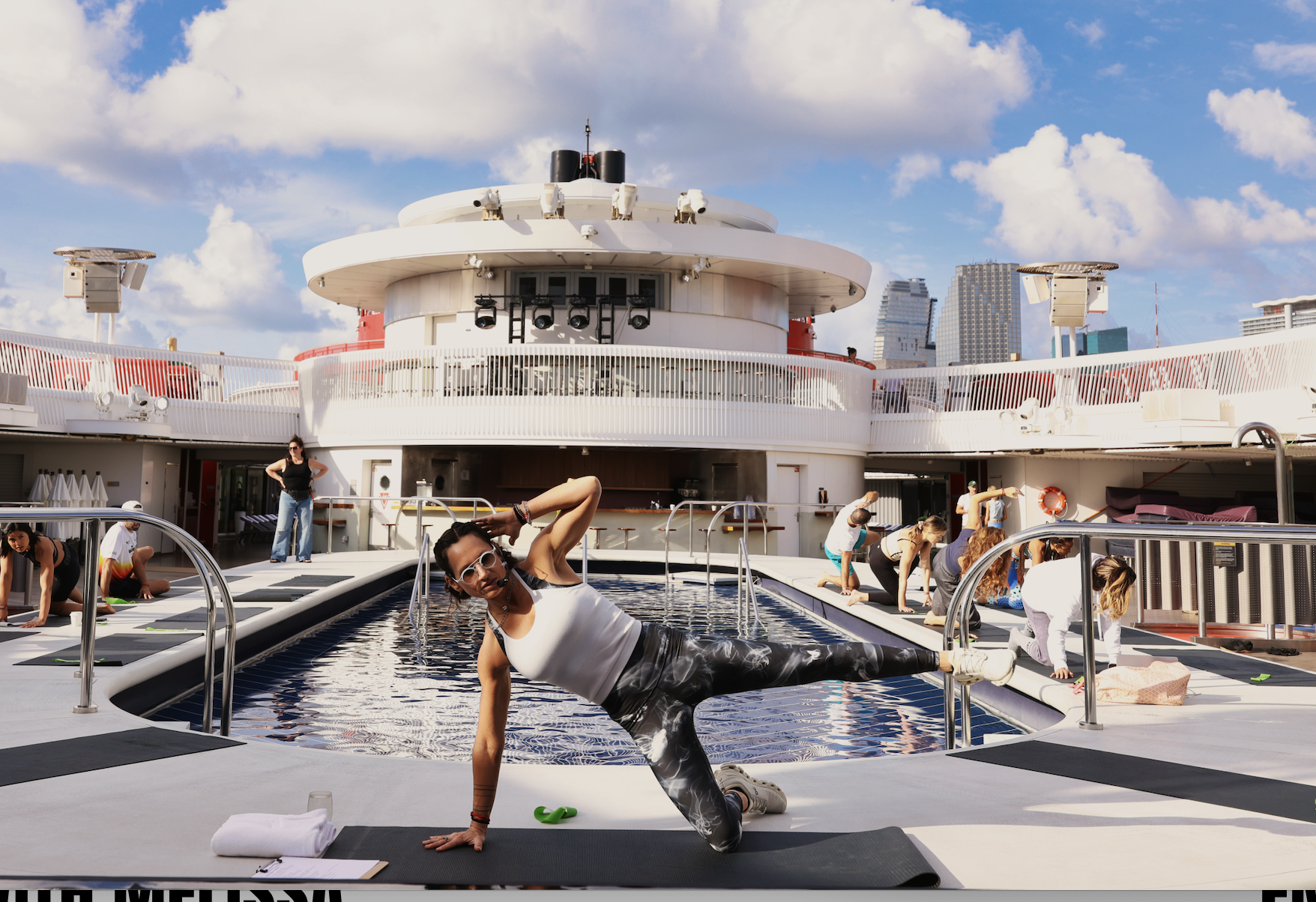People practicing yoga on a pool deck near a cruise ship, with city skyline in the background and a blue sky with clouds.