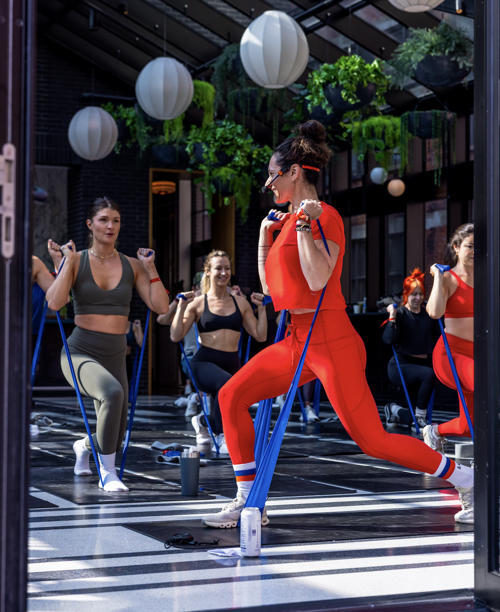 Women participating in an outdoor fitness class using resistance bands, with greenery and decorative lanterns hanging overhead.