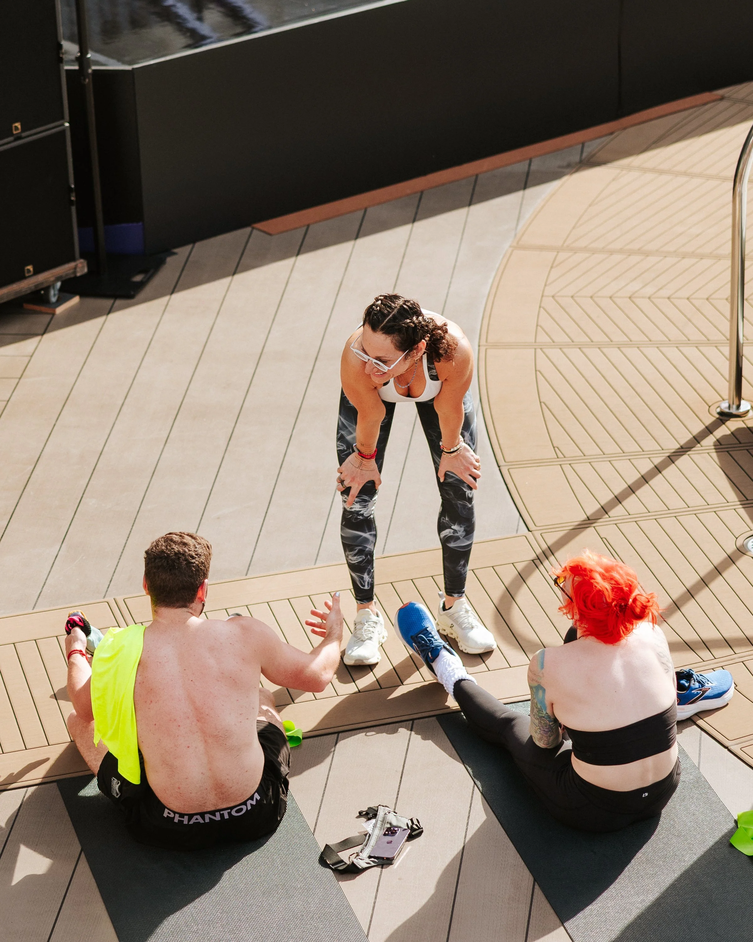 A group of four people engaging in a fitness or stretching session on a rooftop or outdoor area with wooden flooring and exercise mats. The woman standing appears to be instructing or demonstrating a stretch to the seated man and woman.