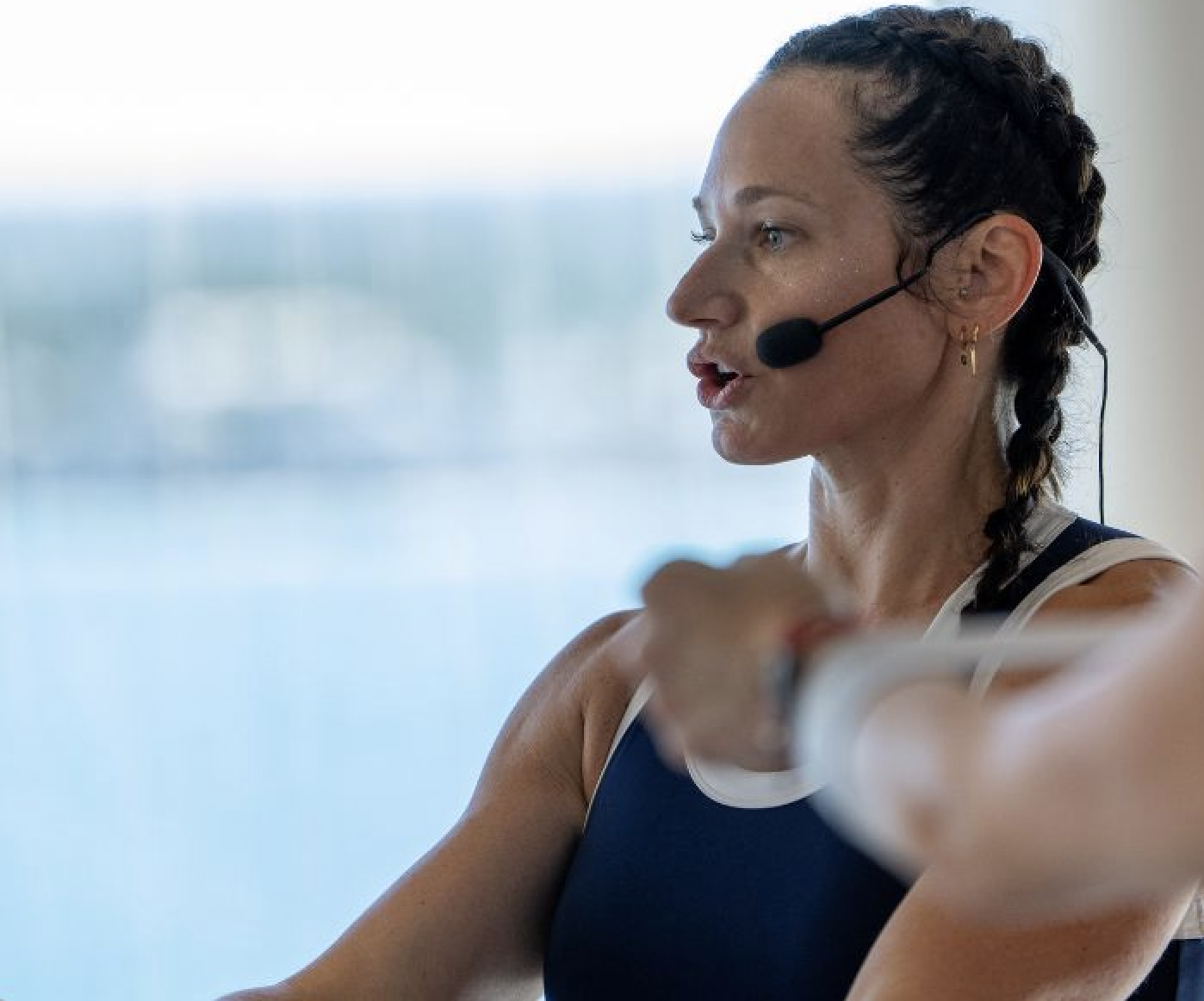 A woman with dark braided hair wearing a headset leads an outdoor fitness class, stretching her arm with a focused expression.