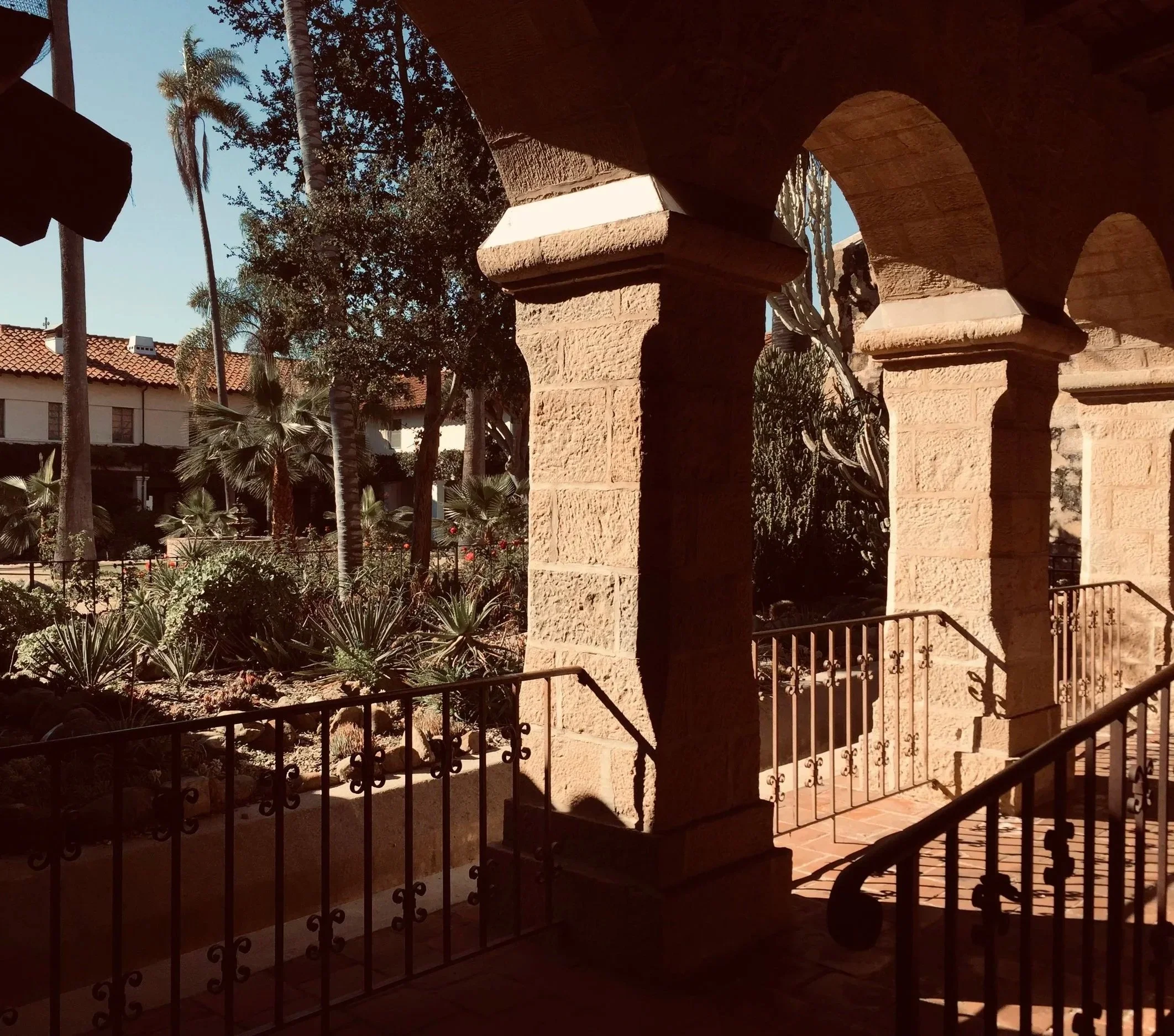 View from a shaded balcony with stone columns, decorative railing, and steps leading down to a garden with palm trees and other plants under a clear blue sky.
