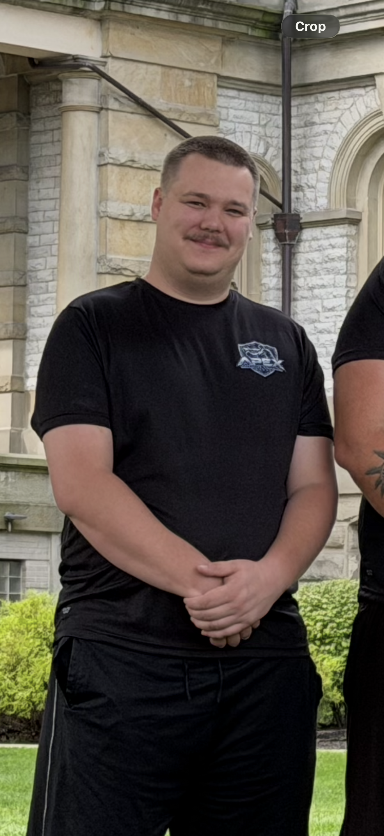 A young man with short hair and a goatee, wearing a black T-shirt with a logo on the chest, standing outdoors in front of a stone building with green bushes in the background.