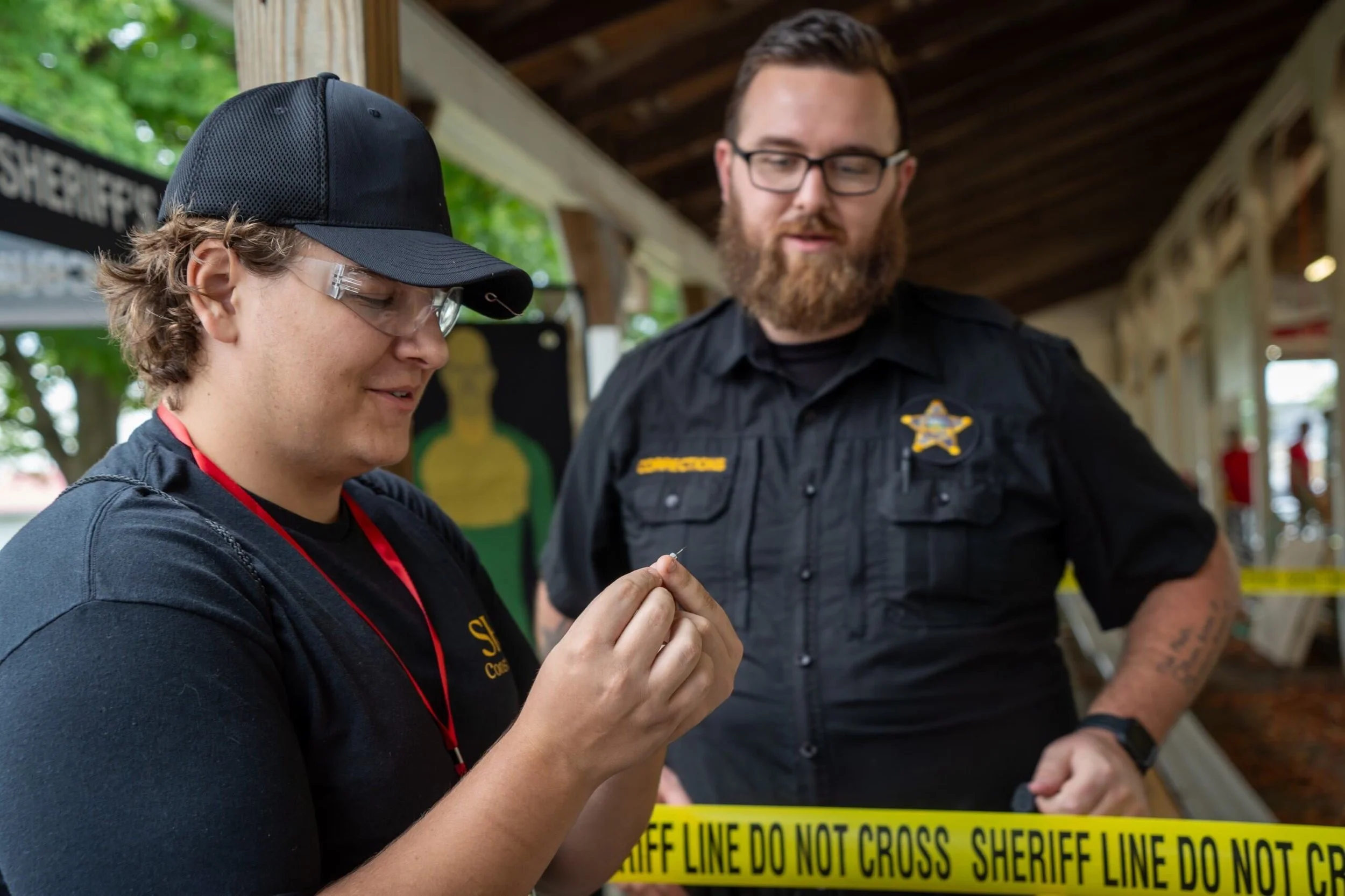 Two law enforcement officers, one wearing glasses and a beard, and the other wearing a black cap and safety glasses, standing behind yellow sheriff line tape that reads 'Sheriff Line Do Not Cross.' They appear to be examining something together indoors, with trees visible outside the open structure.