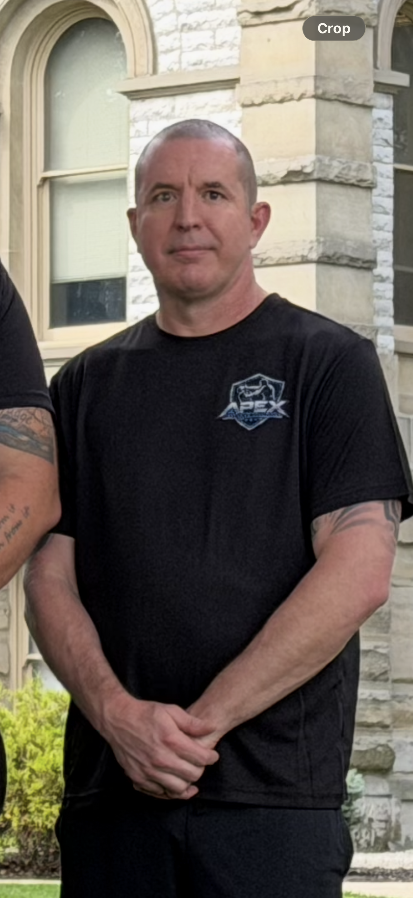 A man standing outdoors in front of a stone building with arched windows, wearing a black T-shirt with a logo on it, with his hands clasped together in front of him.