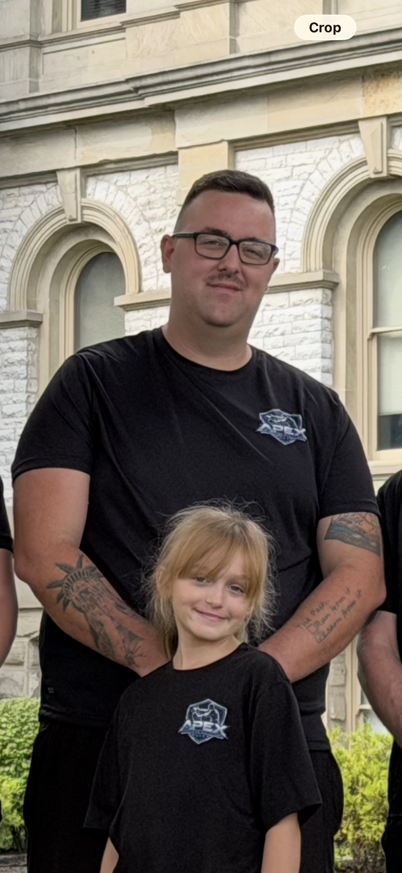 A young girl with blonde hair smiling, standing in front of an adult man with tattoos on his arms, both wearing black t-shirts with a logo. They are outdoors in front of a building with arched windows and white brick walls.