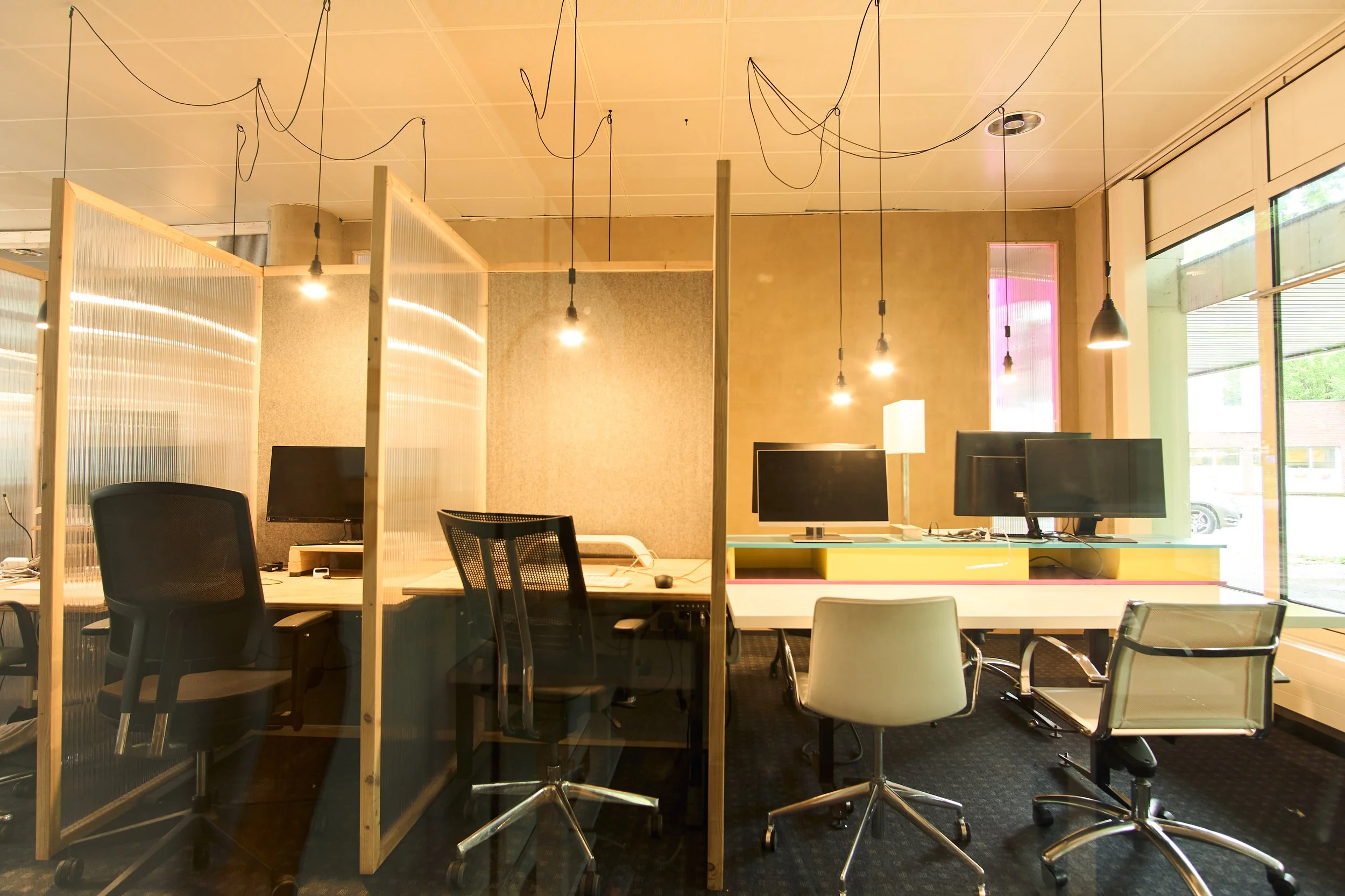 Ein moderner Büroraum mit mehreren Schreibtischen, Computern und ergonomischen Stühlen. Der Raum ist durch Trennwände unterteilt und hat große Fenster mit Blick auf das Freie. Die Decke ist mit Leuchtkörpern und Kabeln versehen.