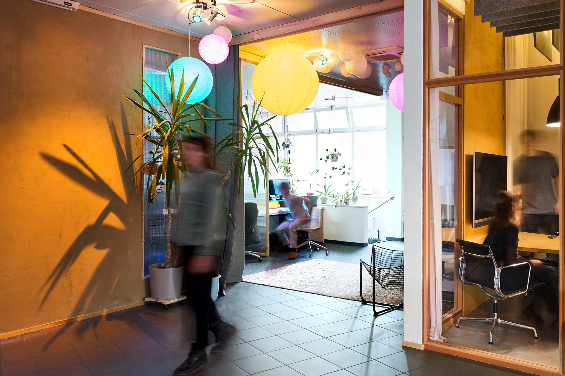 Innenaufnahme eines modernen, lichtdurchfluteten Raumes mit bunten Lampions an der Decke, vielen Pflanzen, einem Schreibtisch mit Computer und mehreren Personen, die sich unterhalten.