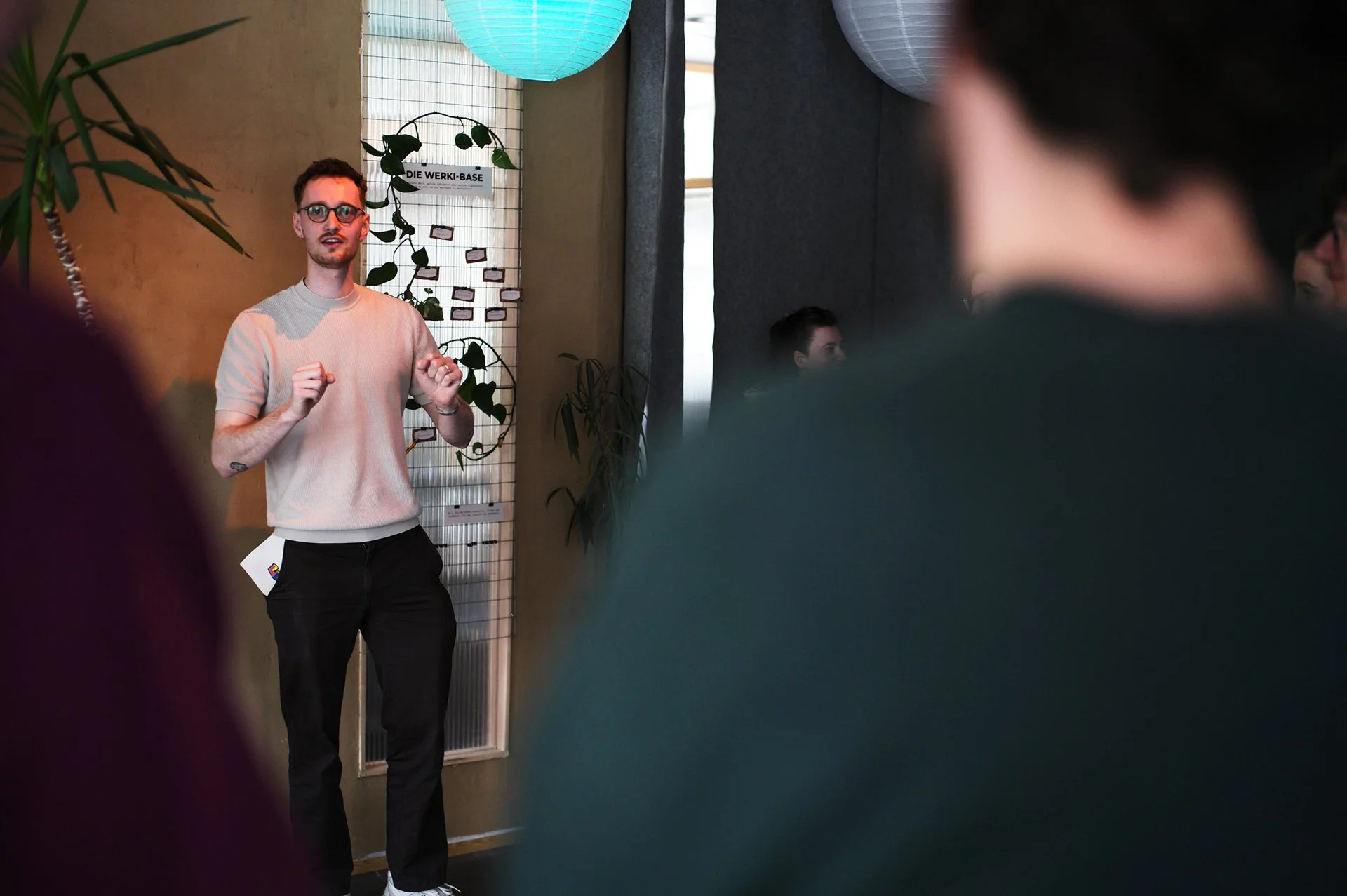 Junge männliche Person mit Brille erklärt something in einer sozialen Umgebung, im Hintergrund Pflanzen und ein Schild mit der Aufschrift "DIE WERK-BASSE"
