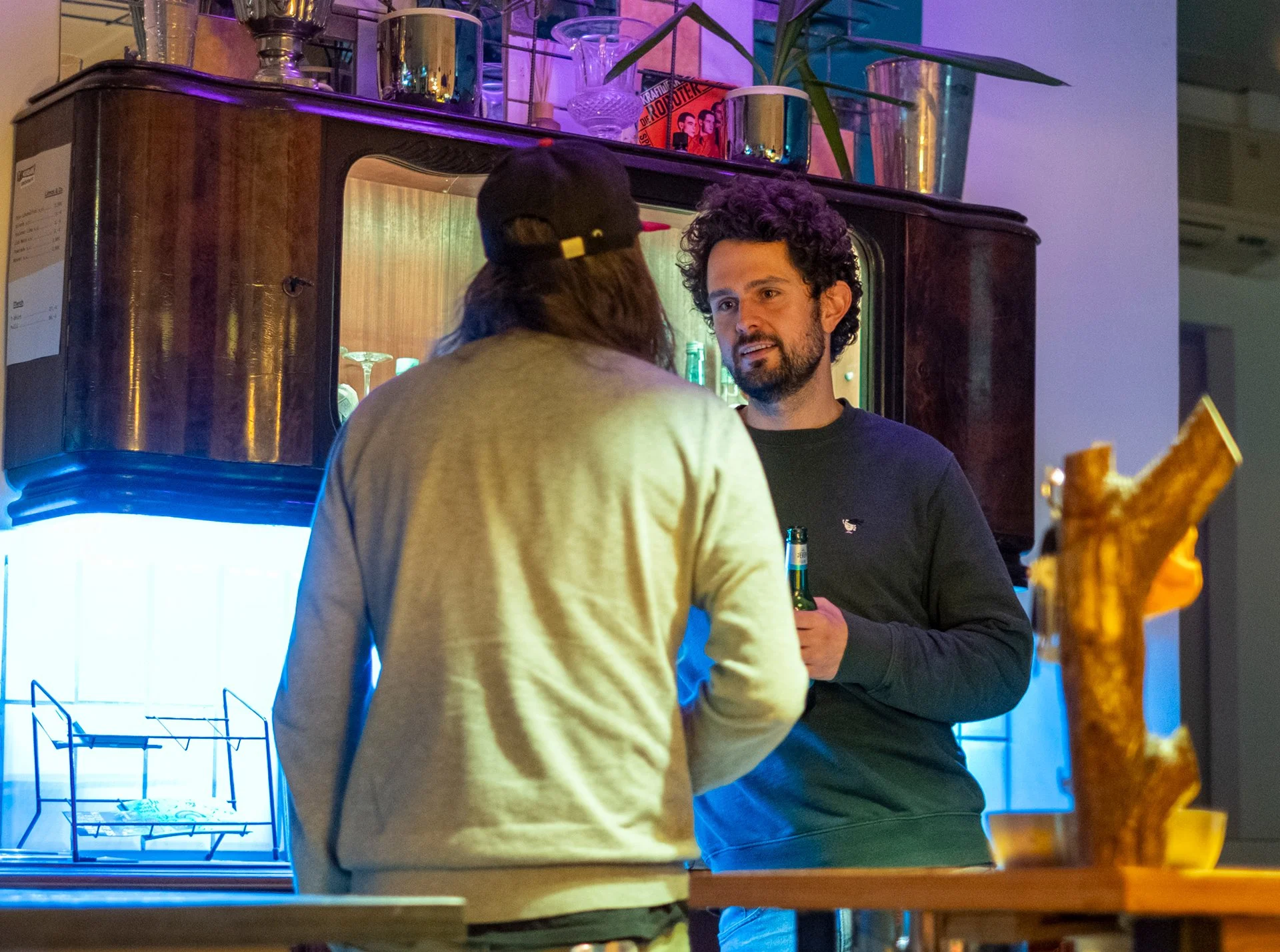 Zwei Männer unterhalten sich an einer Bar im Restaurant. Im Hintergrund sind Gläser, Vasen und Pflanzen auf einem Regal sichtbar.