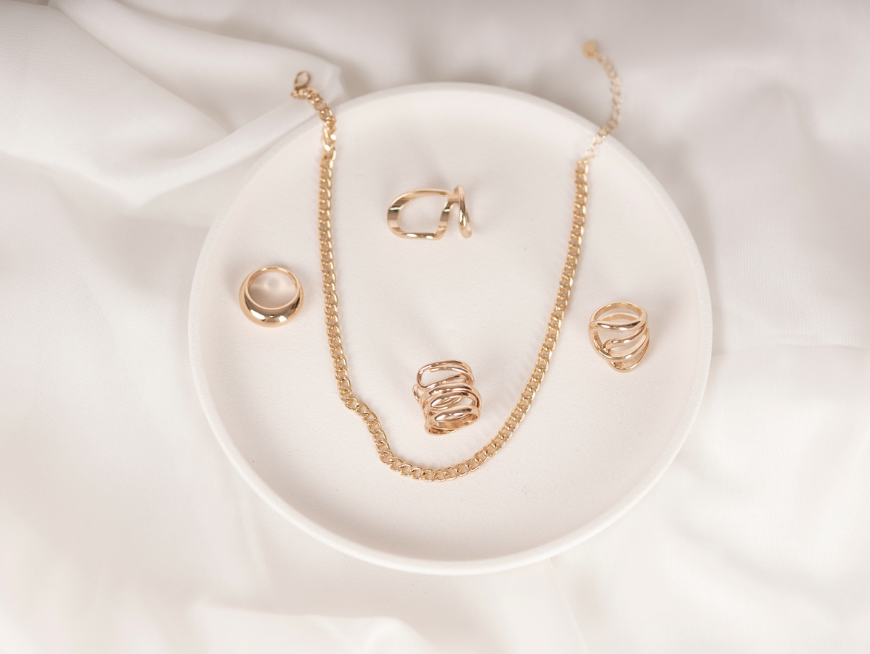 Gold jewelry set including a chain necklace, pair of rings, and a hoop earring displayed on a white dish with a soft fabric background.