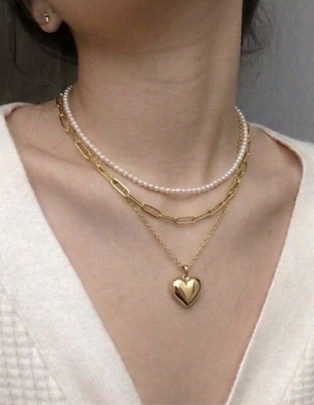 Close-up of a woman's neck and chest displaying three layered necklaces: a pearl choker, a gold chain necklace, and a gold chain with a heart-shaped pendant.
