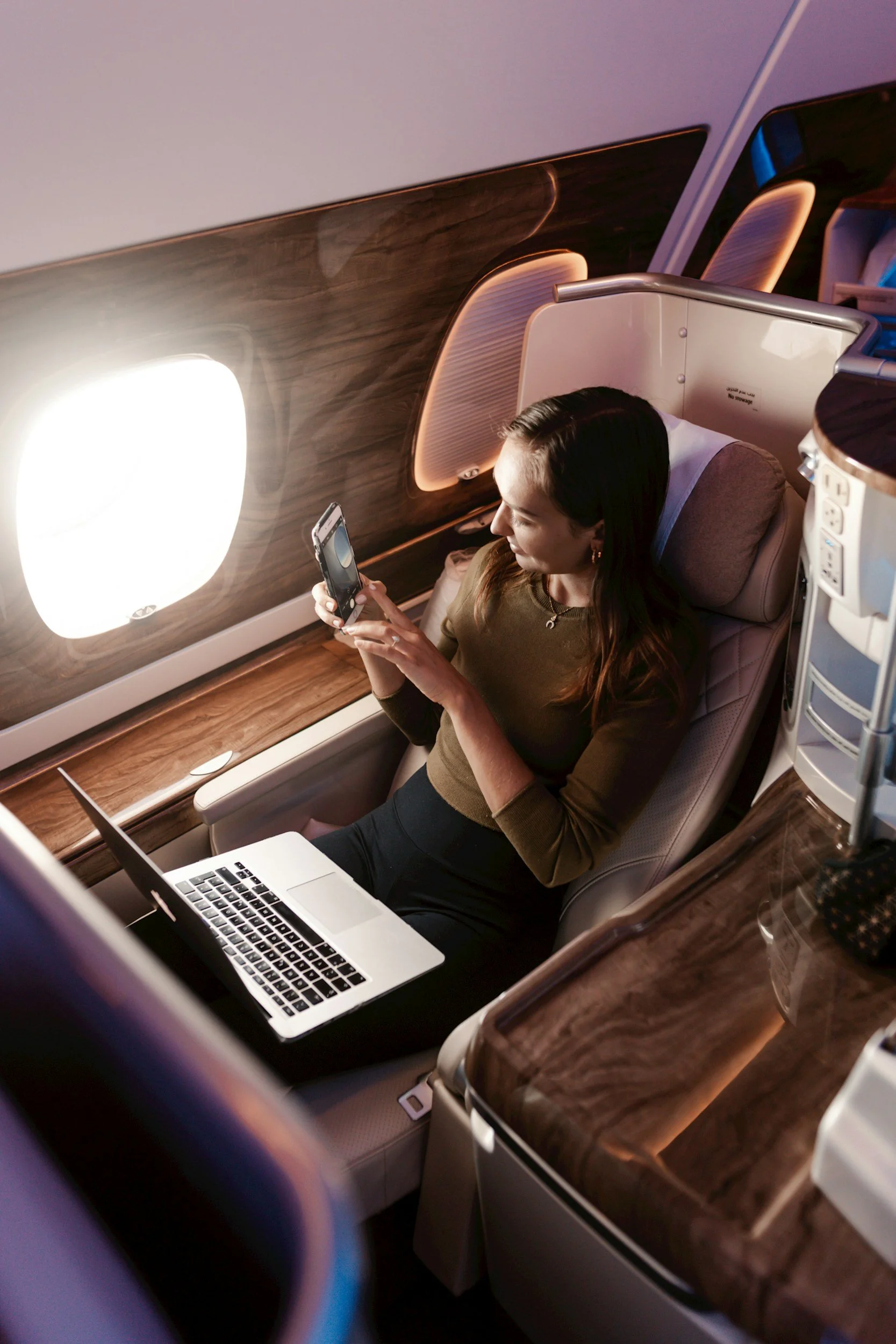 A woman sitting in an airplane seat, looking at her smartphone, with a laptop open on her lap. The airplane window is visible, and the wood-paneled wall adds a warm tone to the cabin interior.