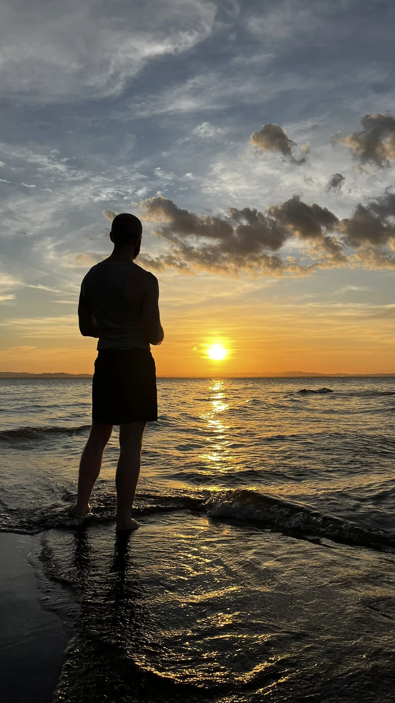 A person standing ankle-deep in the water at the beach during sunset, with a sky filled with clouds and the sun near the horizon.