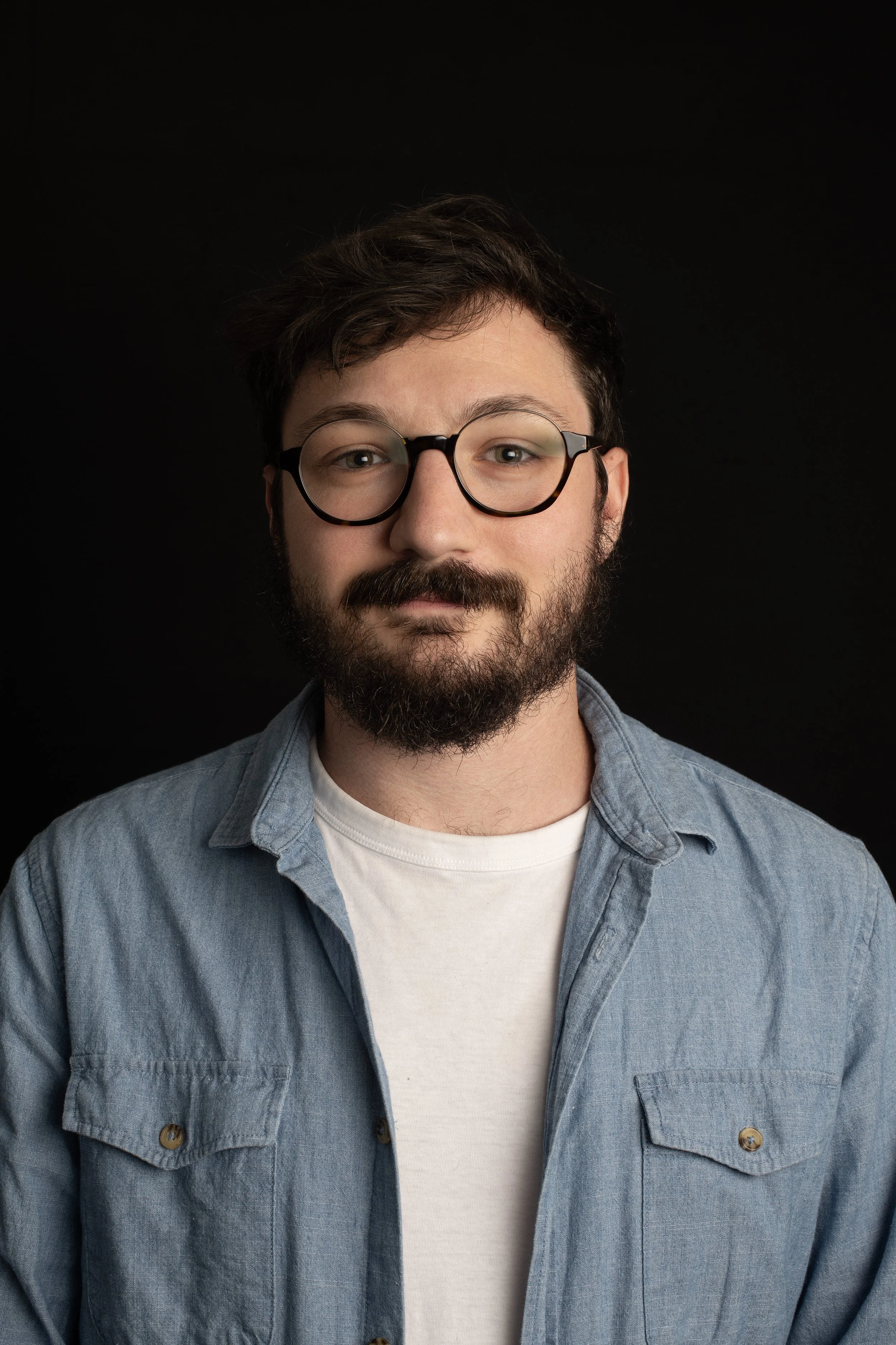 Portrait d'un homme avec lunettes, barbe et cheveux bouclés, portant une chemise en jean et un t-shirt blanc, devant un fond noir.