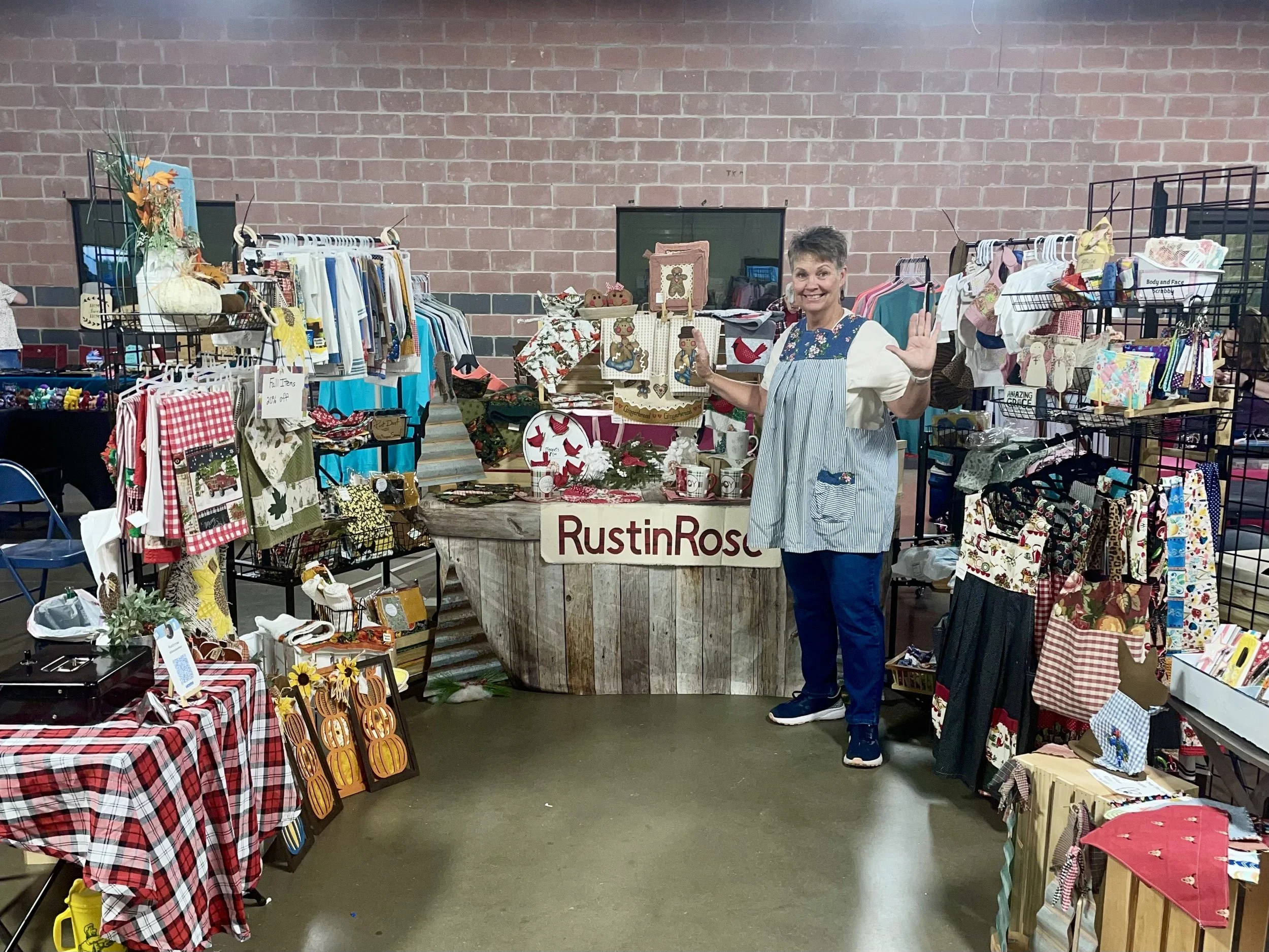 A woman at a craft fair stands behind a display table with Christmas decorations, mugs, and handmade crafts, smiling and waving at the camera.