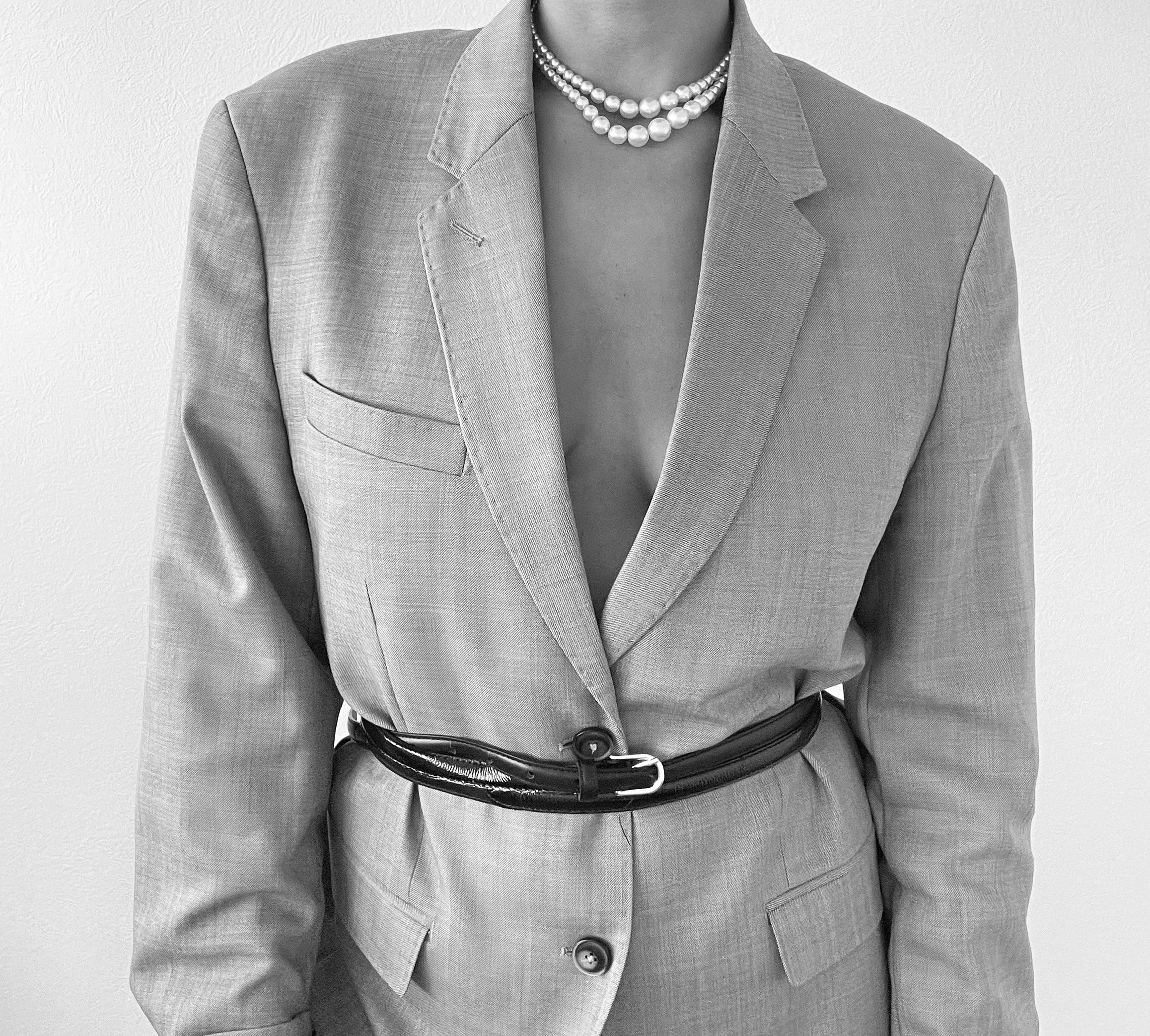 Une personne portant un tailleur gris avec une ceinture noire, un collier de perles et une chemise. L'image est en noir et blanc.