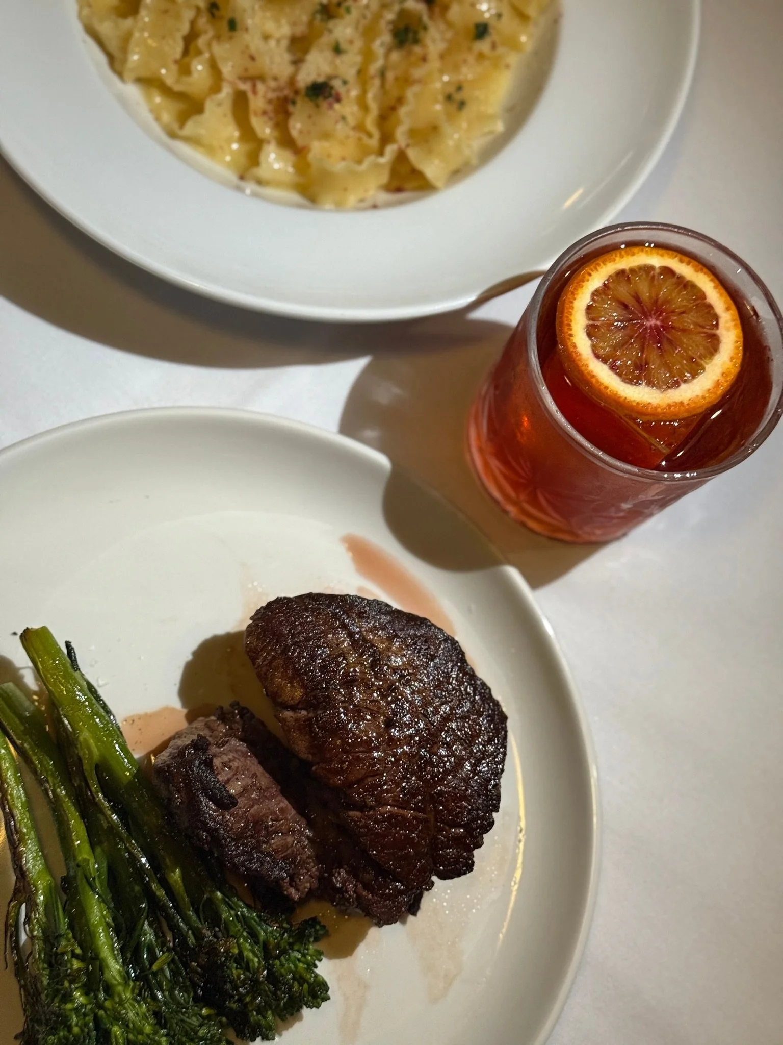 A white plate with two pieces of cooked beef steak and green broccolini, a white bowl with pasta in cream sauce and herbs, and a glass with iced tea and lemon slice on a white table.