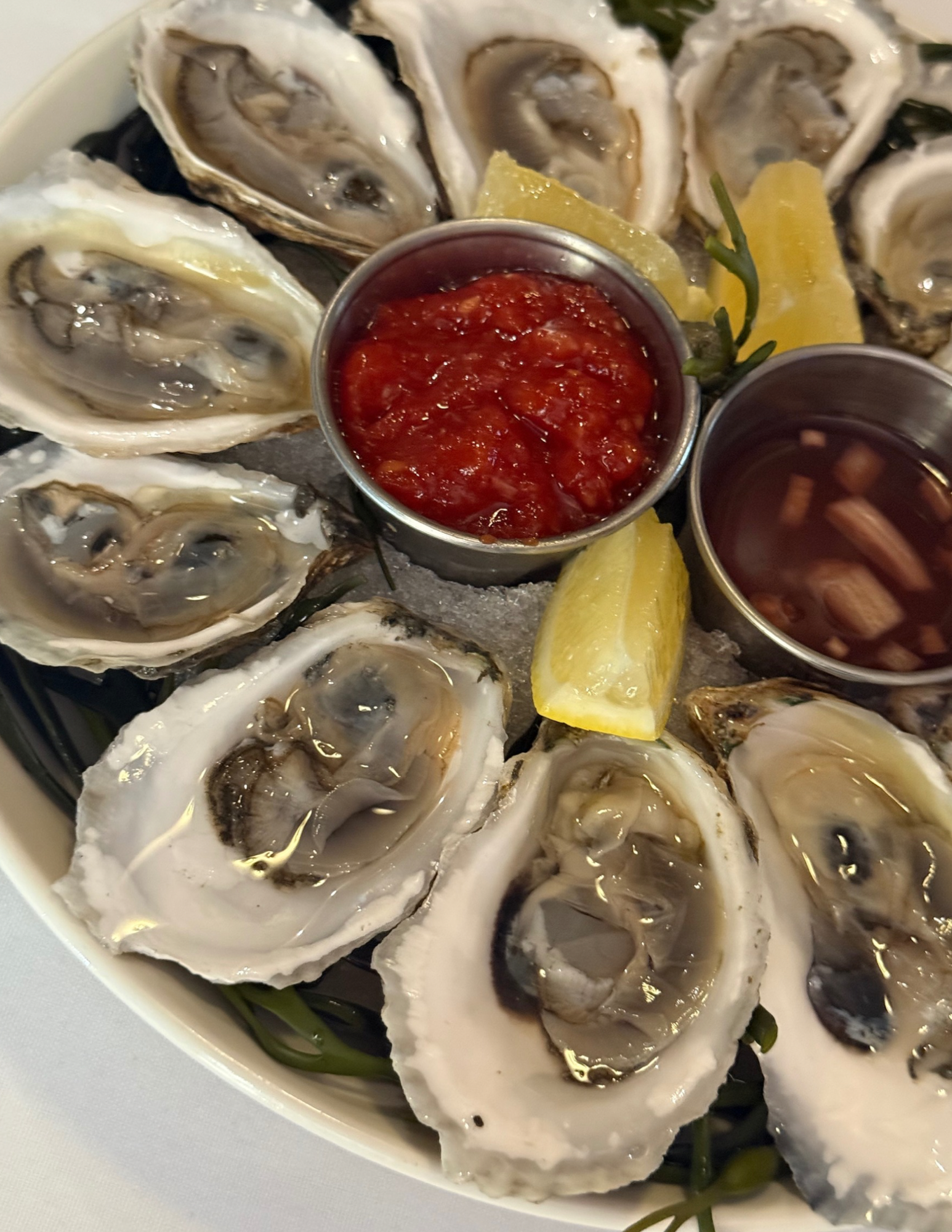 A plate of fresh oysters on the half shell with lemon wedges, cocktail sauce, and mignonette sauce in small metal containers.