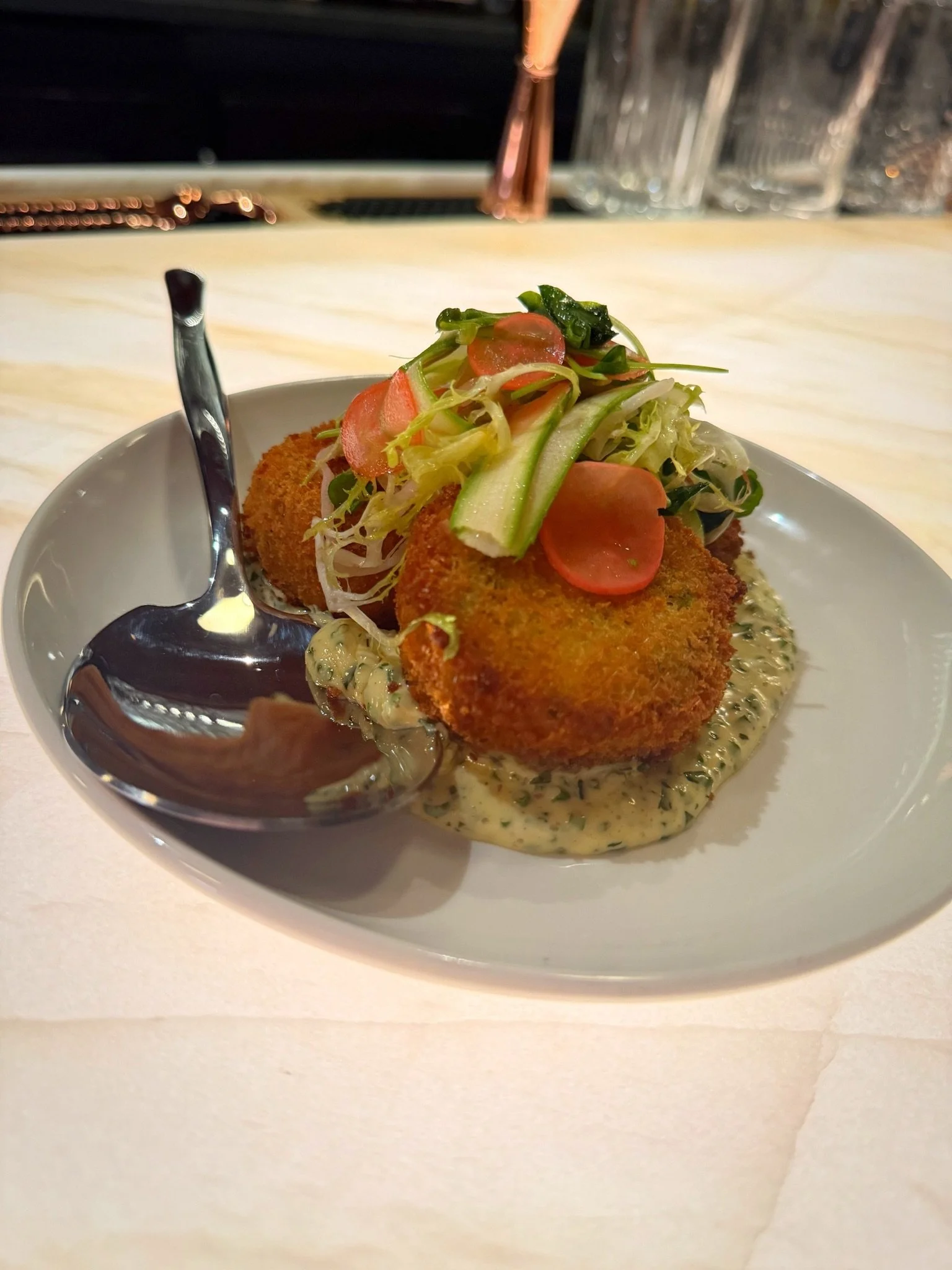 Fried croquettes topped with mixed greens and sliced vegetables, served on a bed of creamy sauce in a white bowl with a spoon.