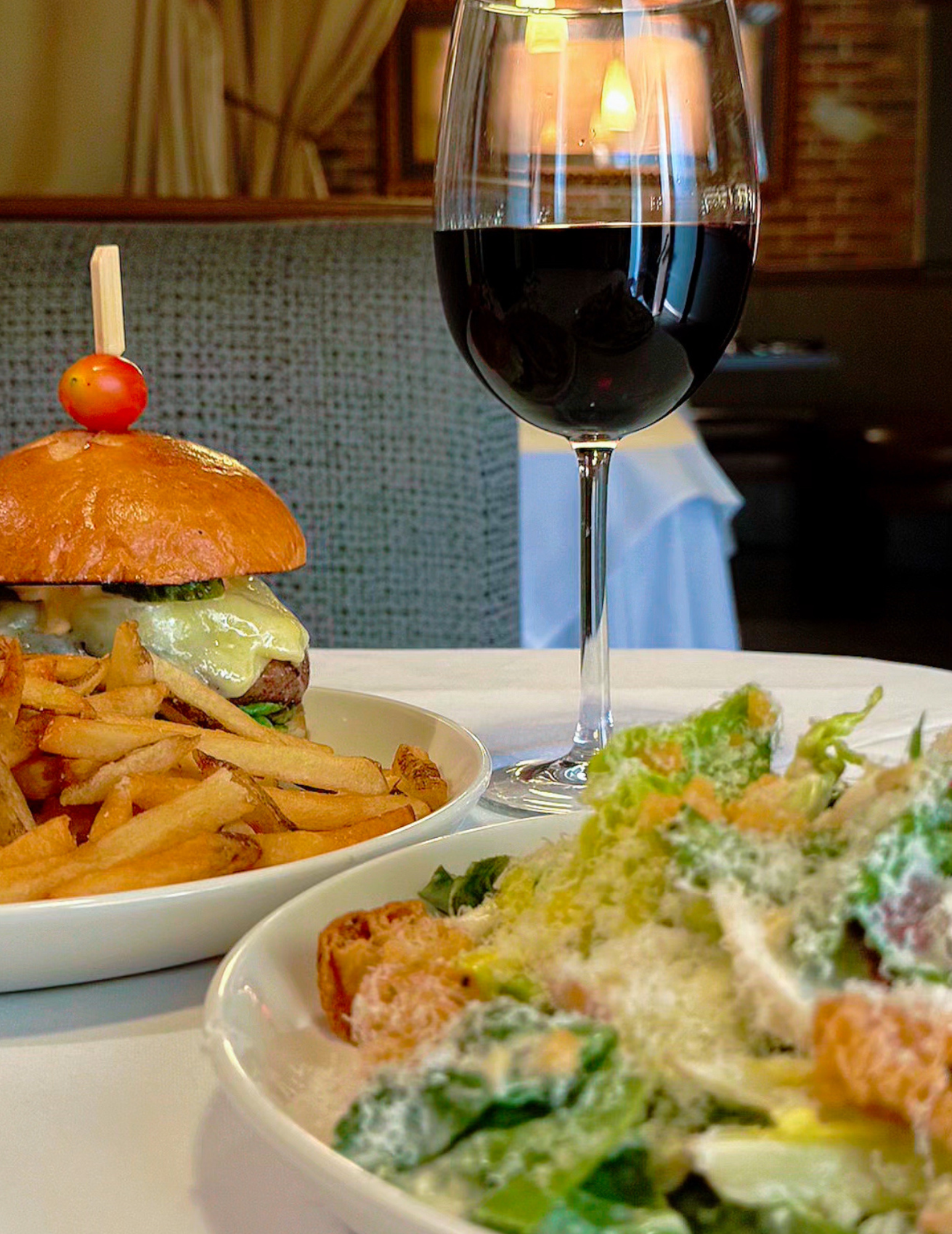 A table with a plate of fries, a burger with lettuce, tomato, cheese, and a cherry tomato inserted with a toothpick, a glass of red wine, and a Caesar salad with croutons, lettuce, and grated cheese in a dining setting.