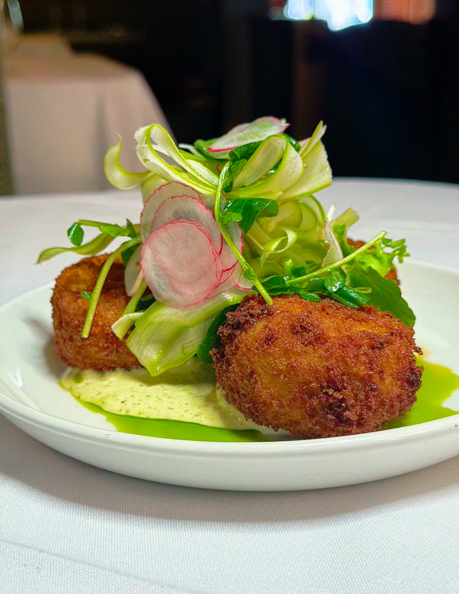 Fried croquettes topped with fresh microgreens and thinly sliced radishes, served with a green sauce and a creamy yellow sauce on a white plate.