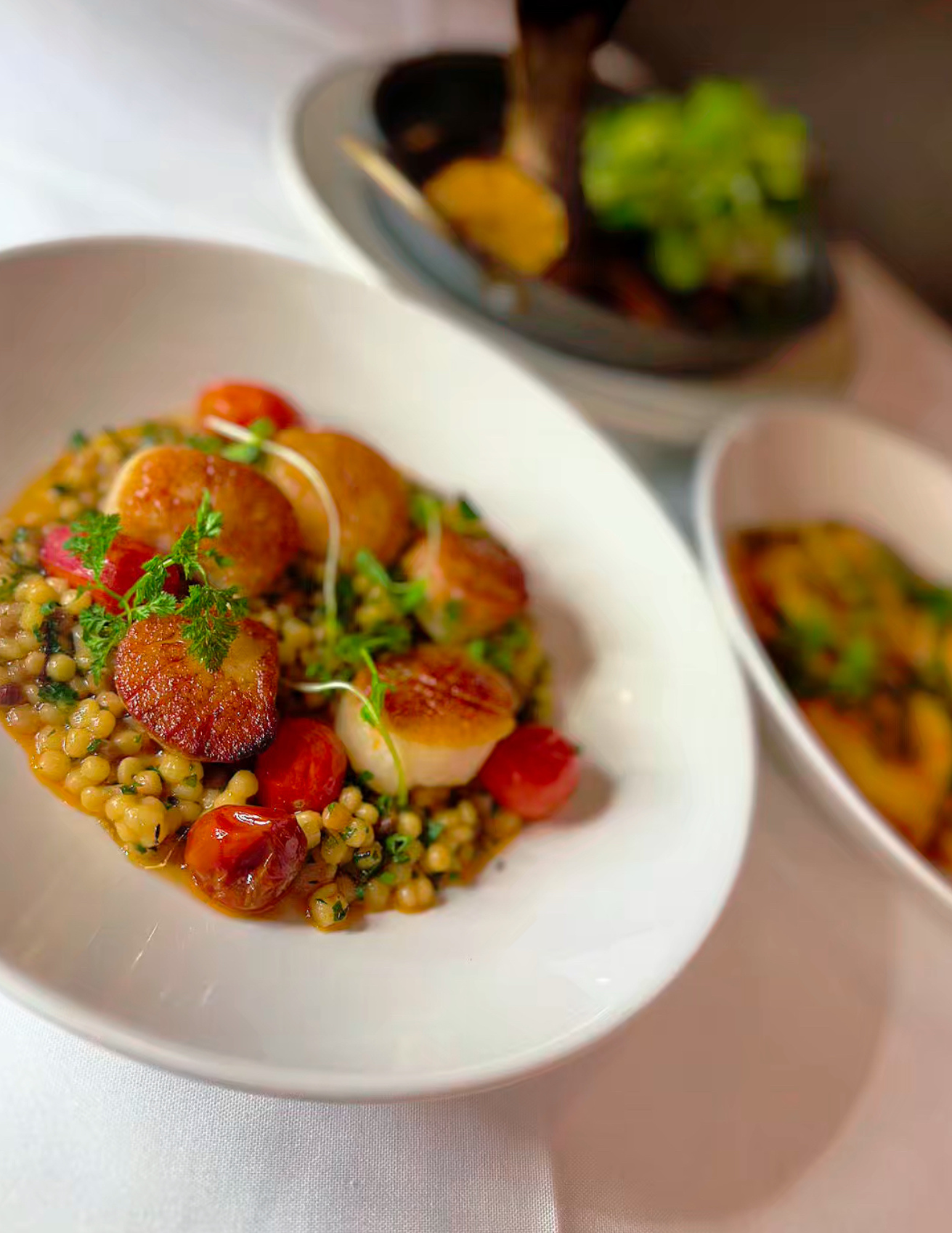 Close-up of a plated dish with grains, berries, and green herbs, along with two small bowls of soup or sauce in the background.