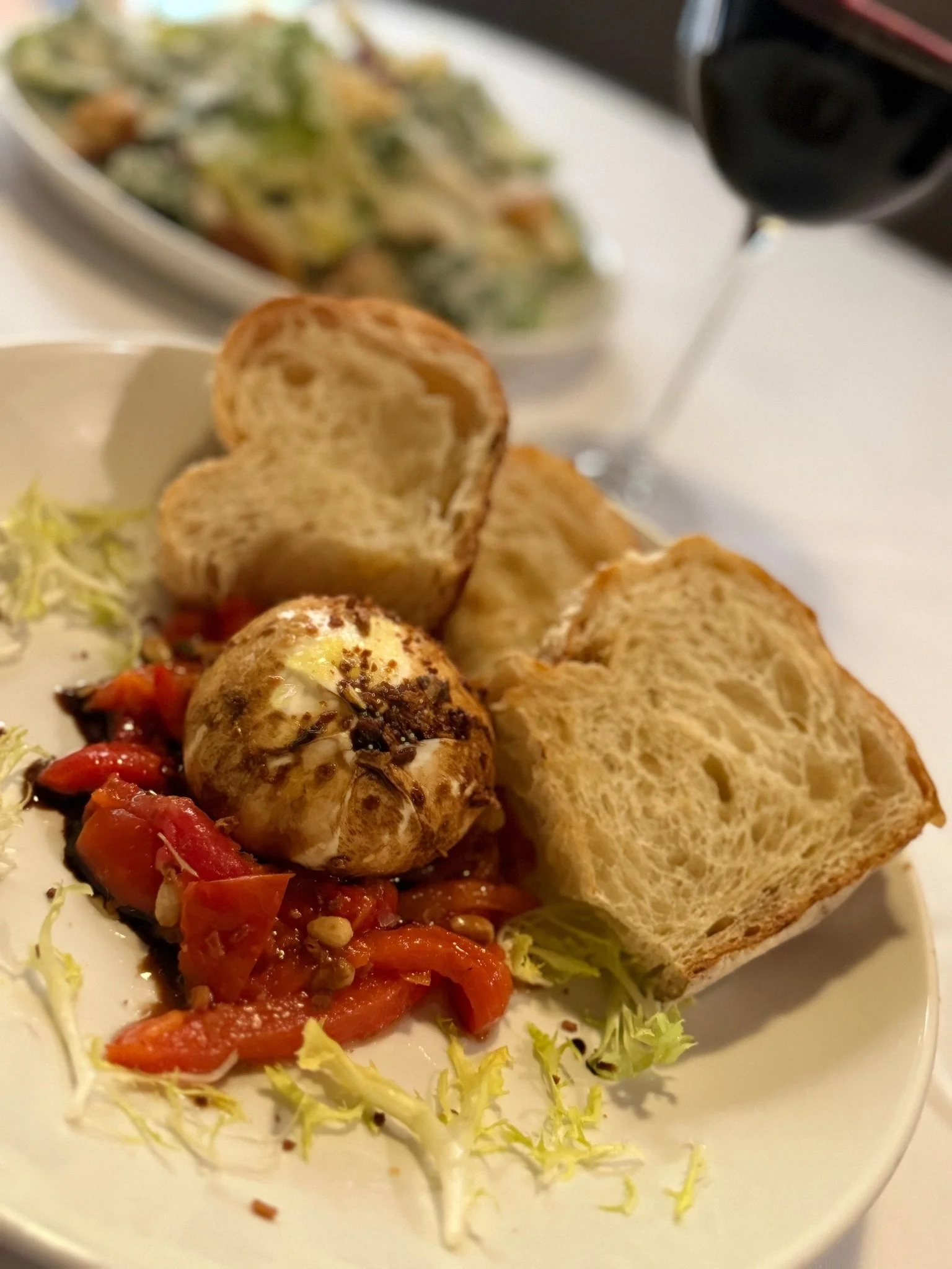 A plate of roasted eggplant topped with tomato and chili sauce, served with toasted bread slices, beside a small amount of lettuce, and a glass of red wine in the background.