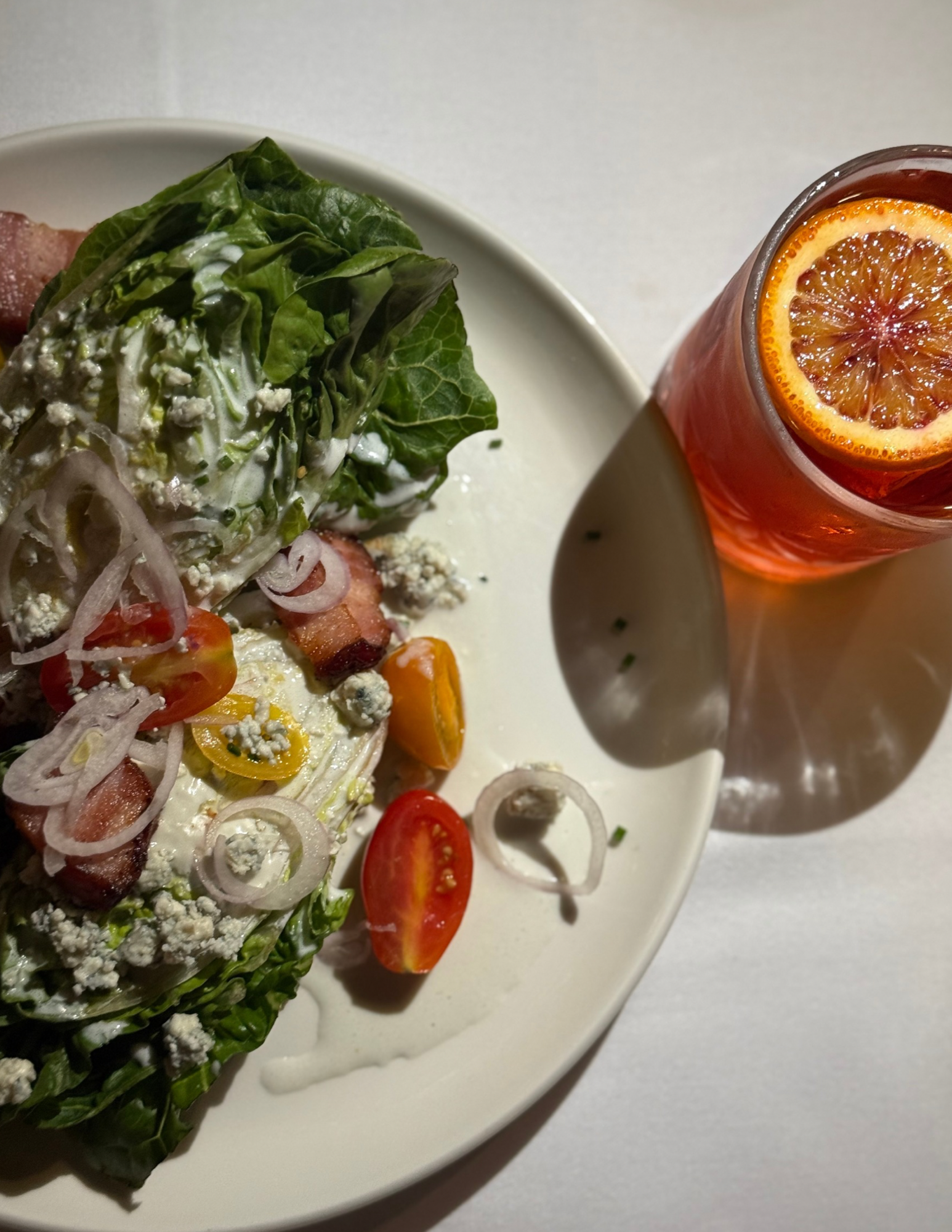 Salad on a white plate with cherry tomatoes, onions, bacon, and salad greens, served with a side of red soda with a sliced citrus fruit in it.