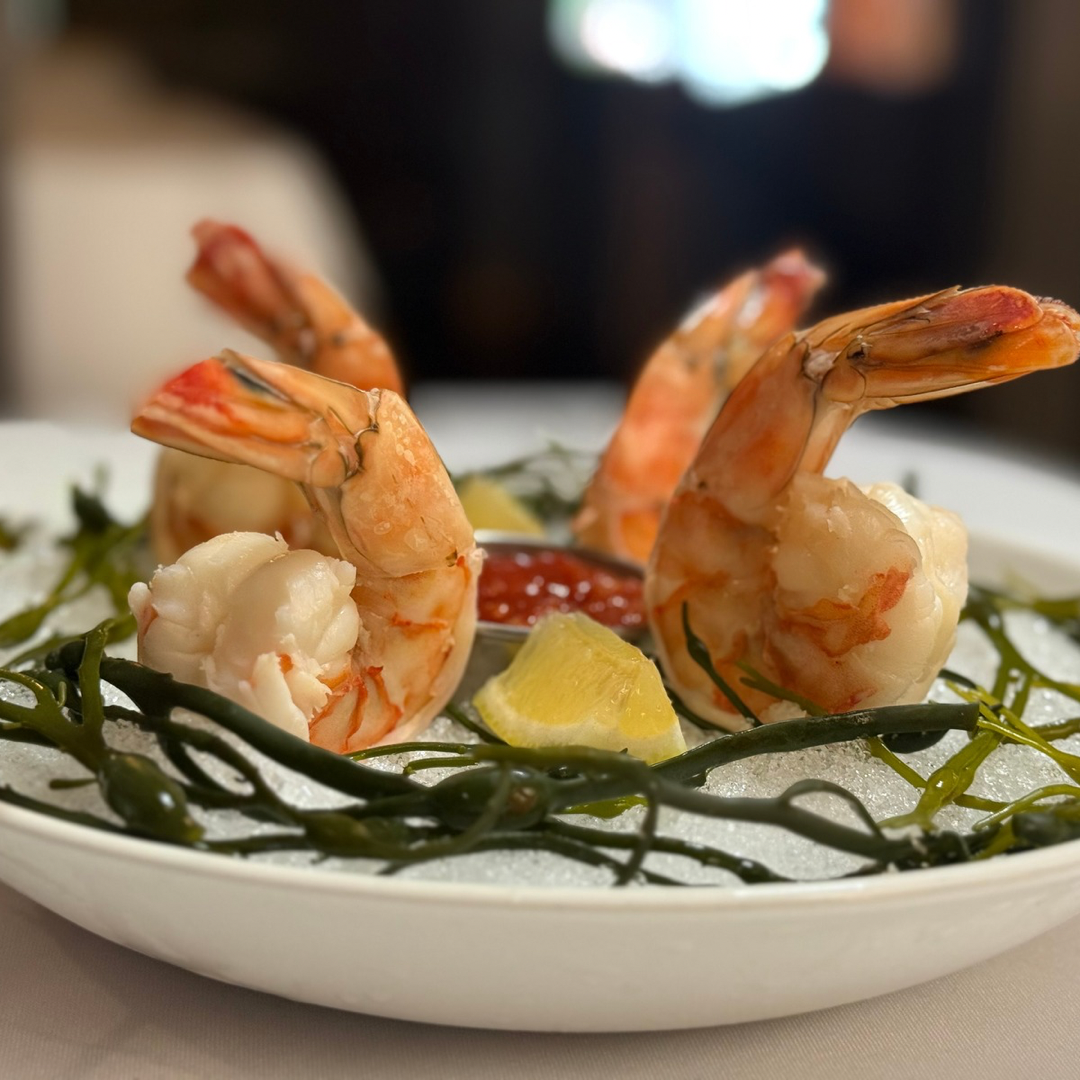 Seafood dish with shrimp, lemon wedges, seaweed, and a small bowl of red sauce, served on a bed of ice.