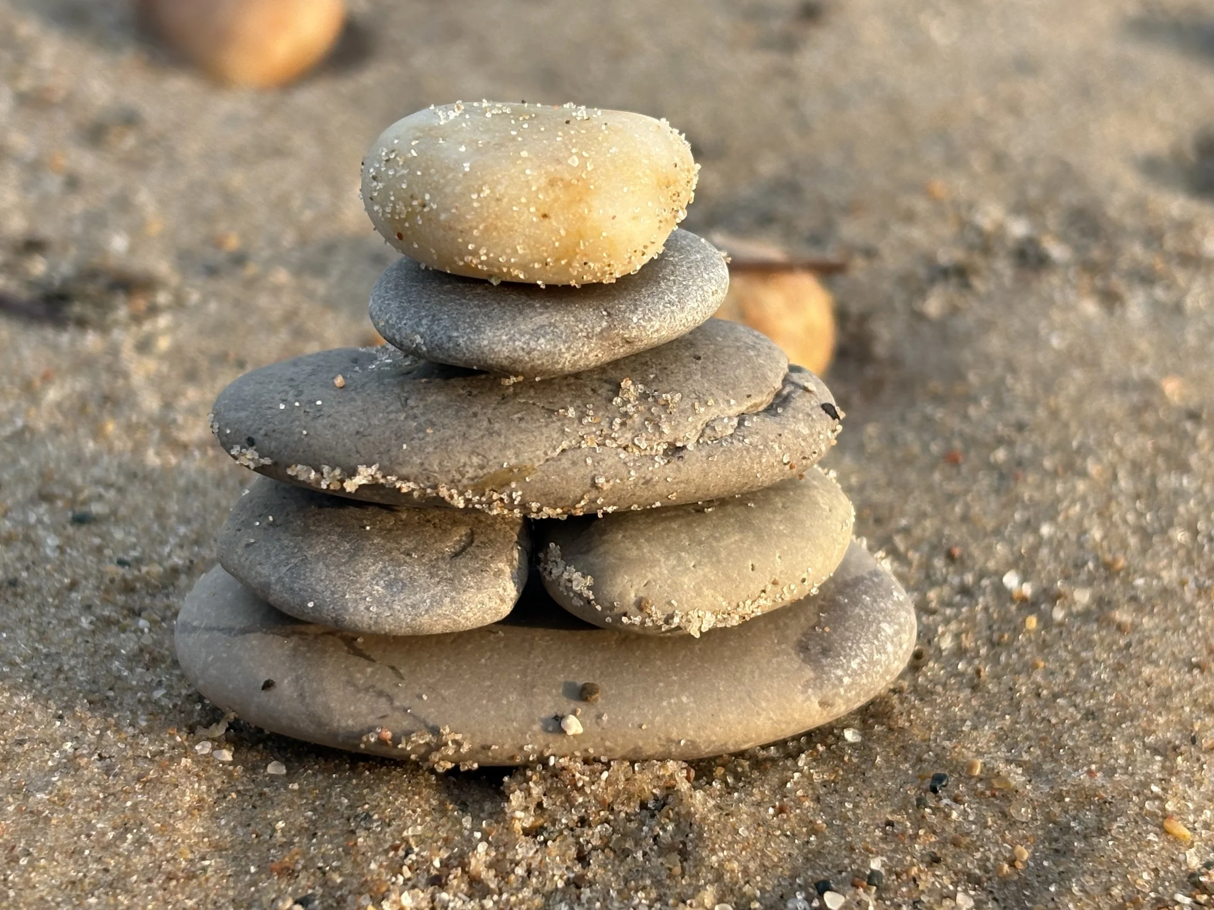 A stack of five smooth, flat stones balanced on top of each other on a sandy beach, with the sand and other stones in the background.