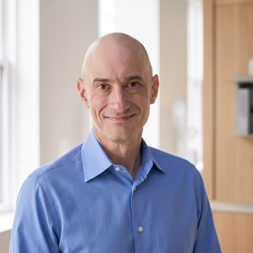 A bald man in a blue dress shirt standing indoors in front of a window and bookshelf, smiling at the camera.