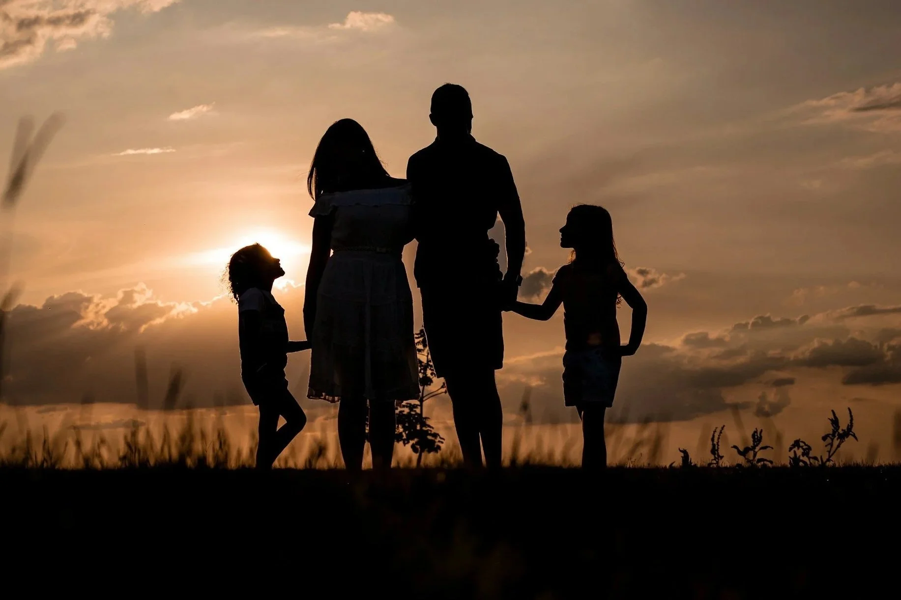 Famiglia di quattro persone, due adulti e due bambini, che si tengono per mano e si trovano all'aperto al tramonto, con cielo nuvoloso sullo sfondo.