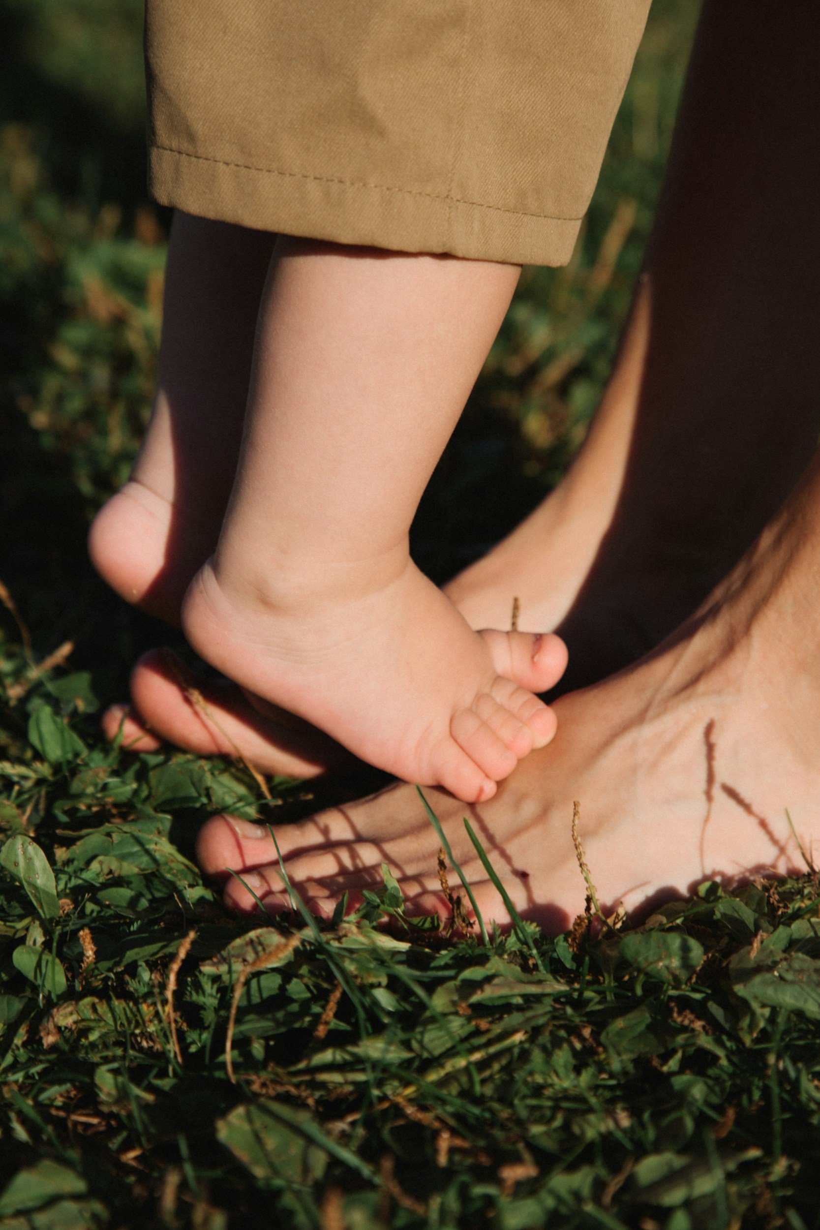 Un bambino si tiene per mano con un adulto, tutti e due sono sdraiati sull'erba, con il bambino che tocca il suolo con il piede e la manina dell'adulto. L'attenzione è sulle loro mani e sui piedi, con uno sfondo di erba verde.